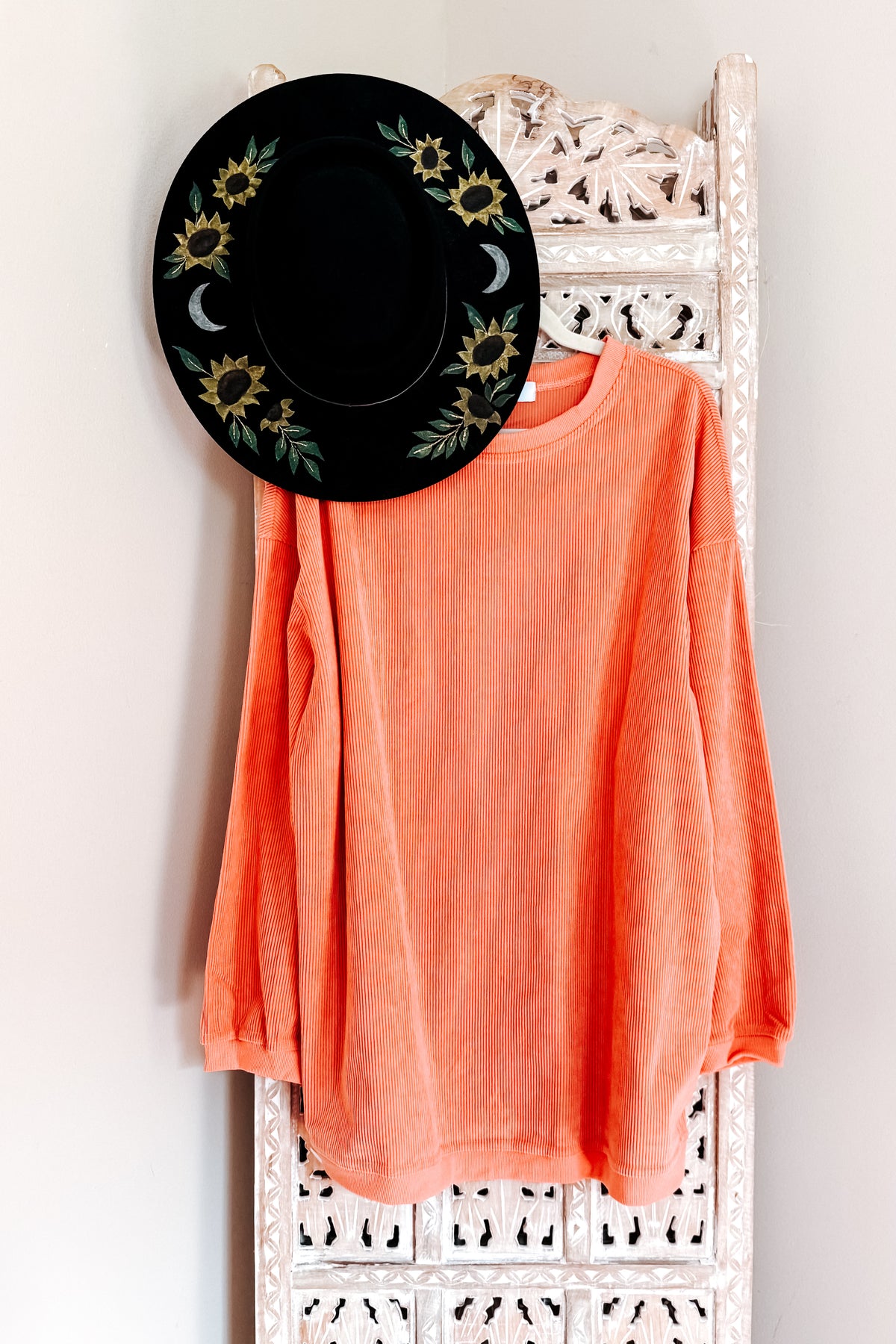Orange oversized corded sweatshirt with a ribbed round neckline and drop shoulders hanging on an ornate white rack beneath a black floral hat
