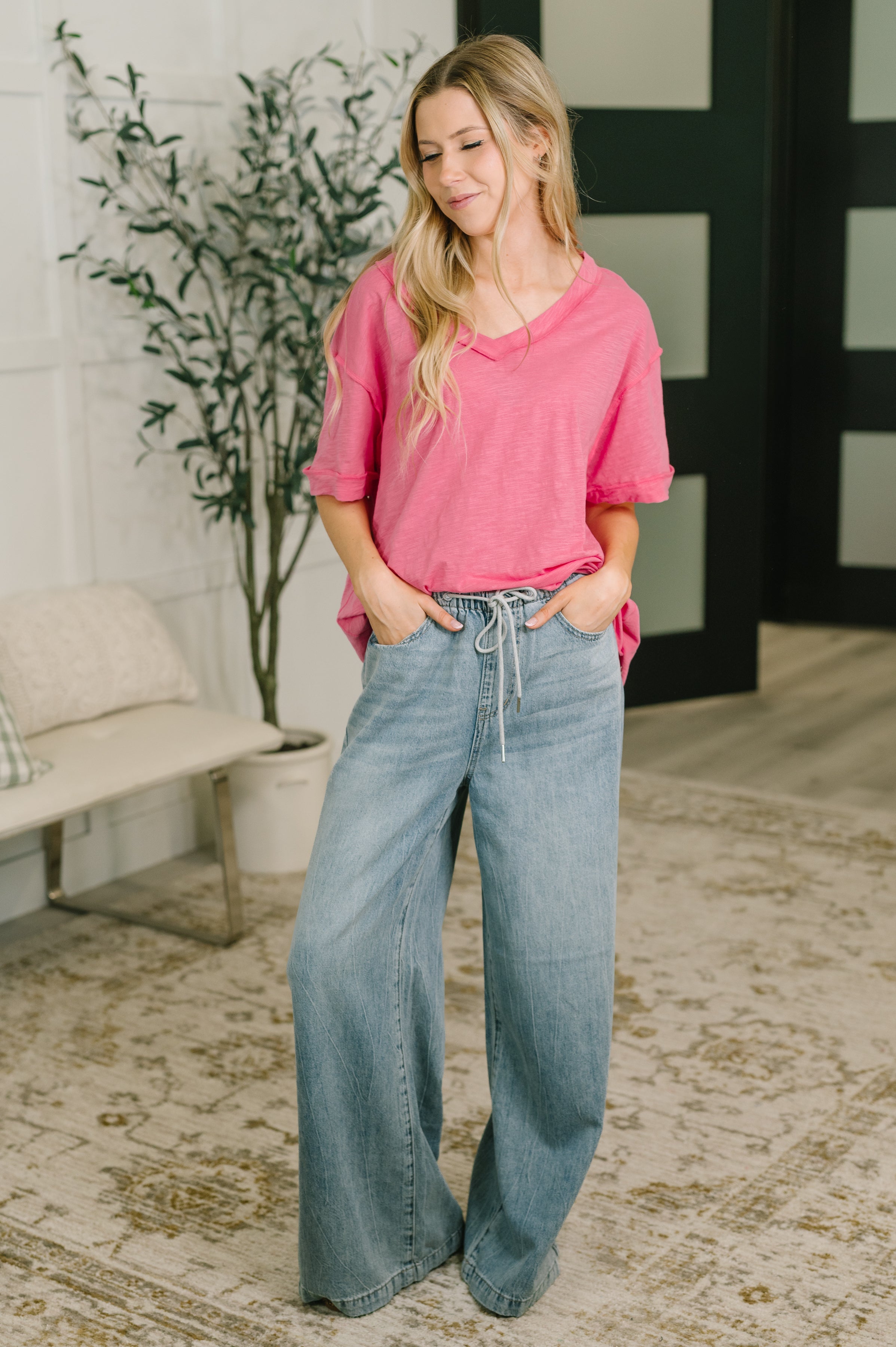 Woman stands indoors wearing relaxed pink V-neck tee paired with wide-leg light wash jeans in casual setting