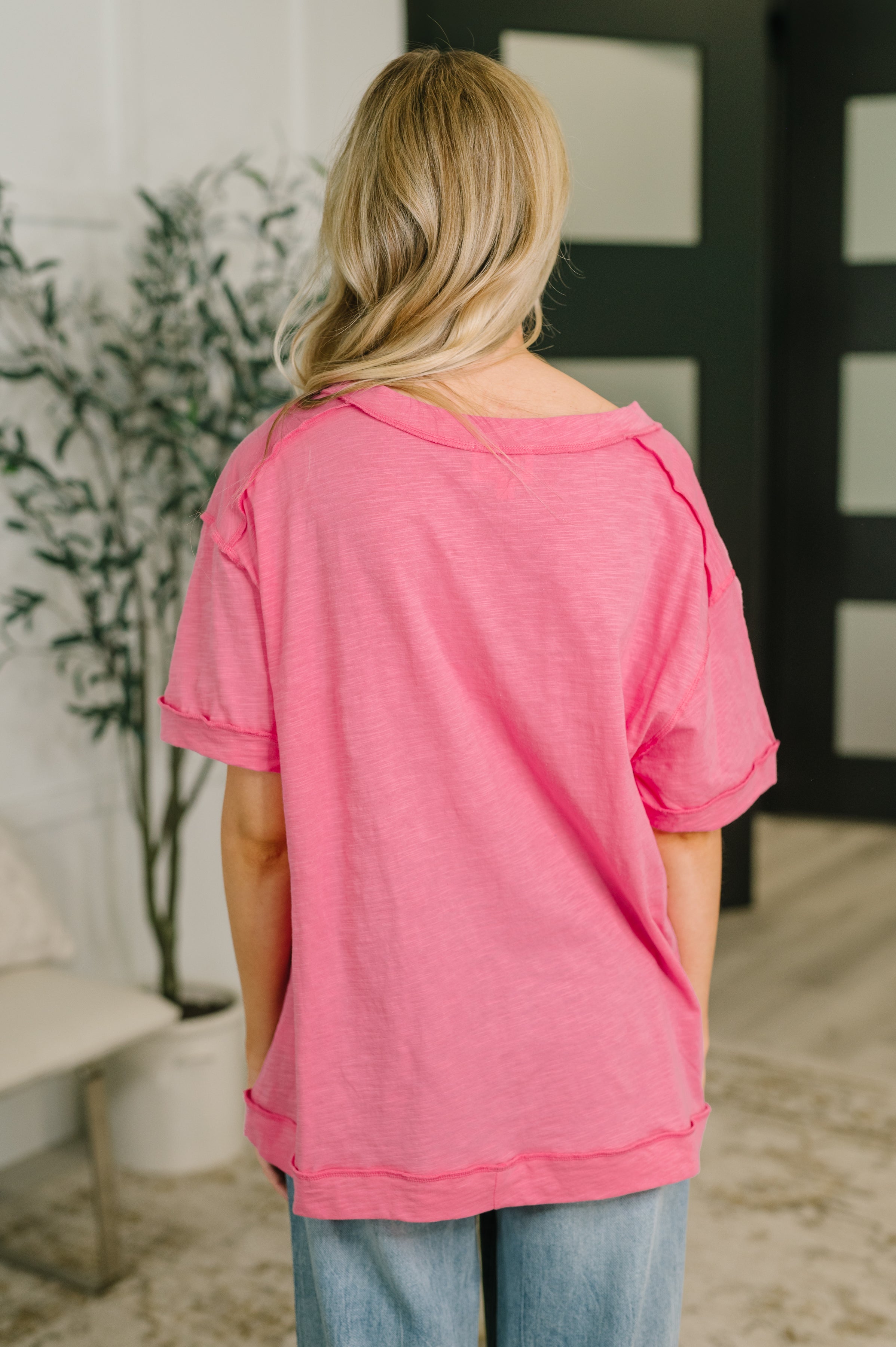 Back view of woman wearing pink relaxed fit tee with rolled sleeves in casual indoor setting with plant and modern decor