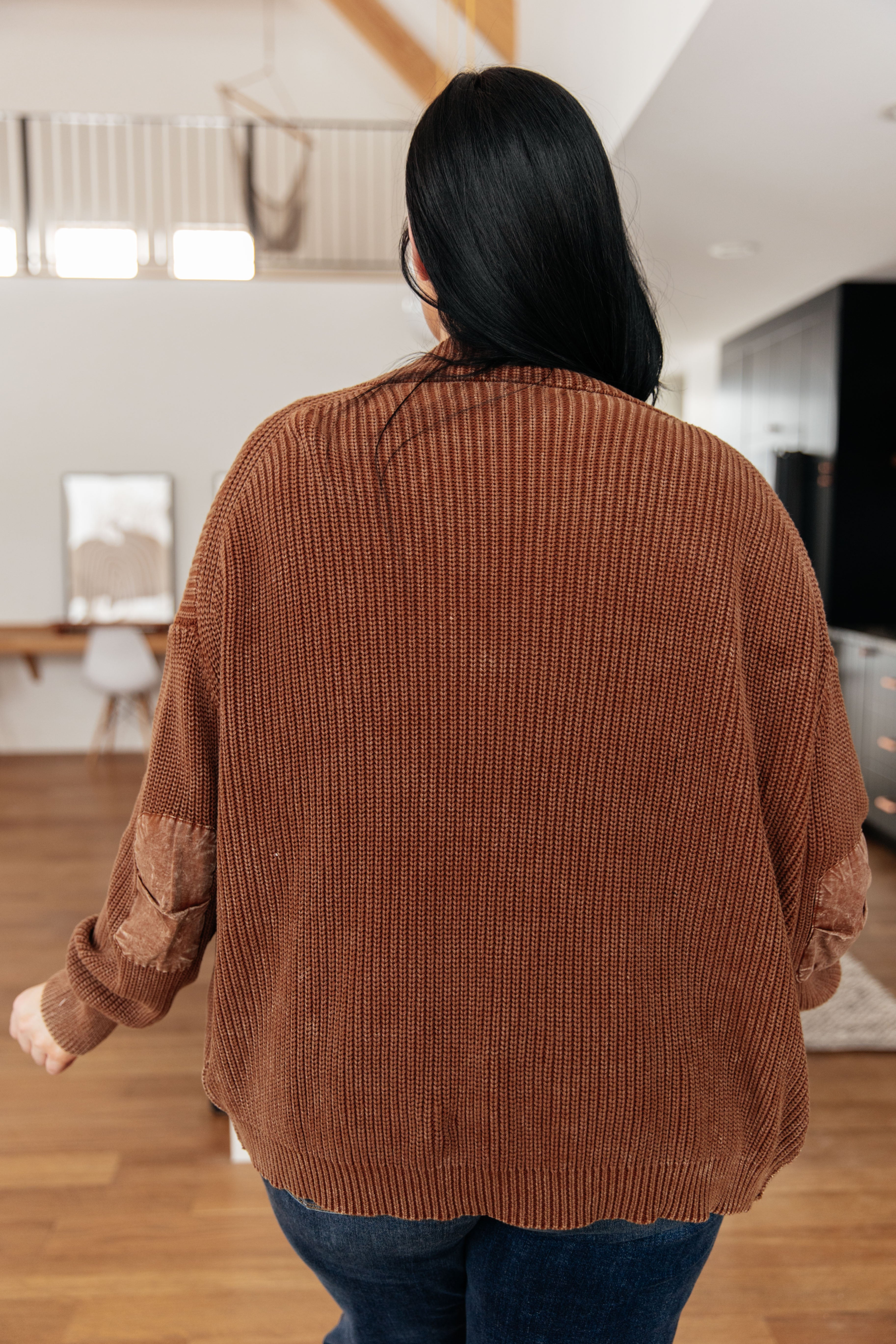 Woman wearing a rust brown chunky knit cardigan with velvet elbow patches standing in a modern home interior