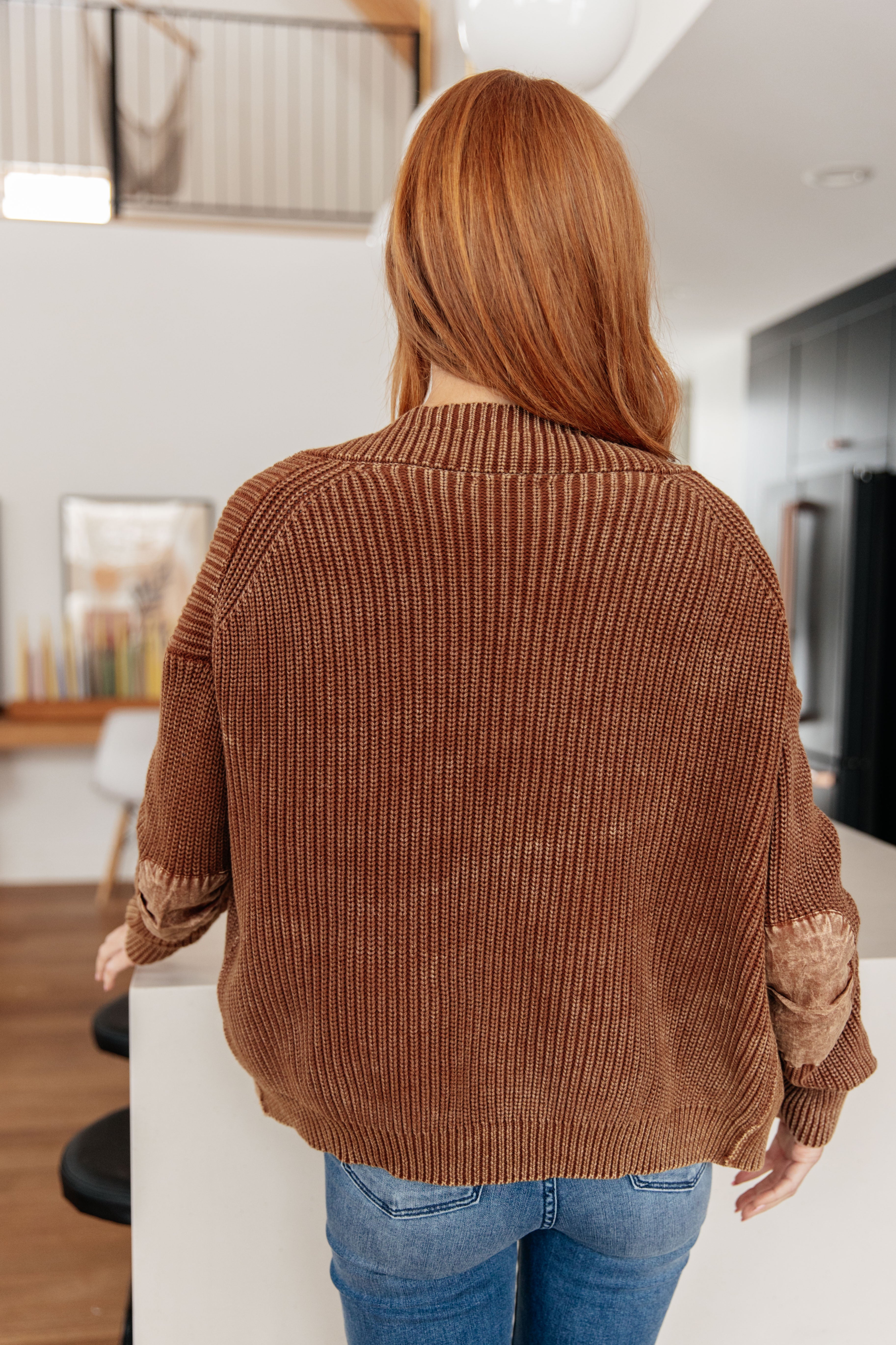Woman wearing a brown chunky knit cardigan with patch details and blue jeans in a bright modern kitchen setting