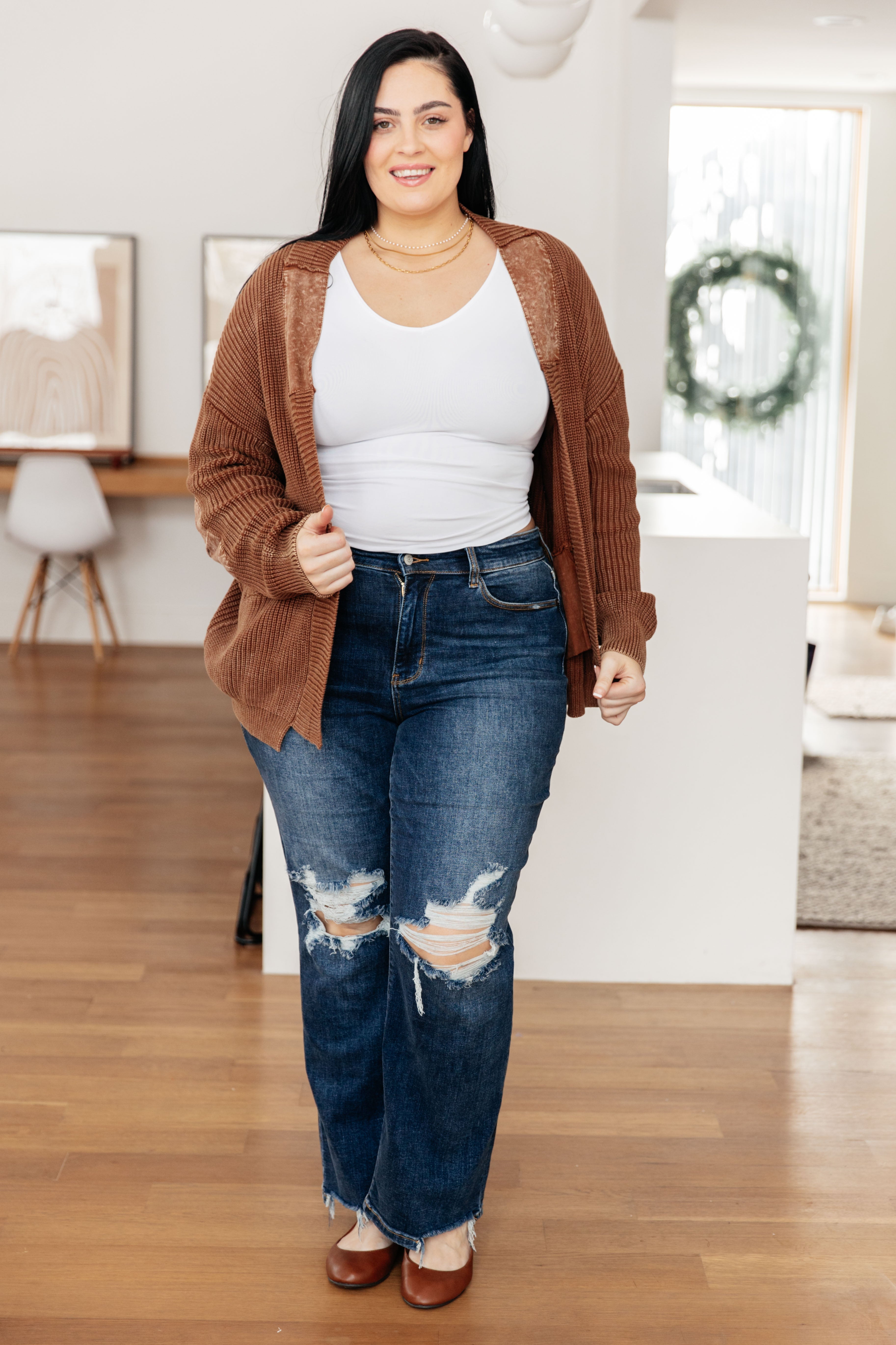 Curvy woman wearing a brown knit cardigan over white tank top paired with ripped blue jeans and brown shoes indoors