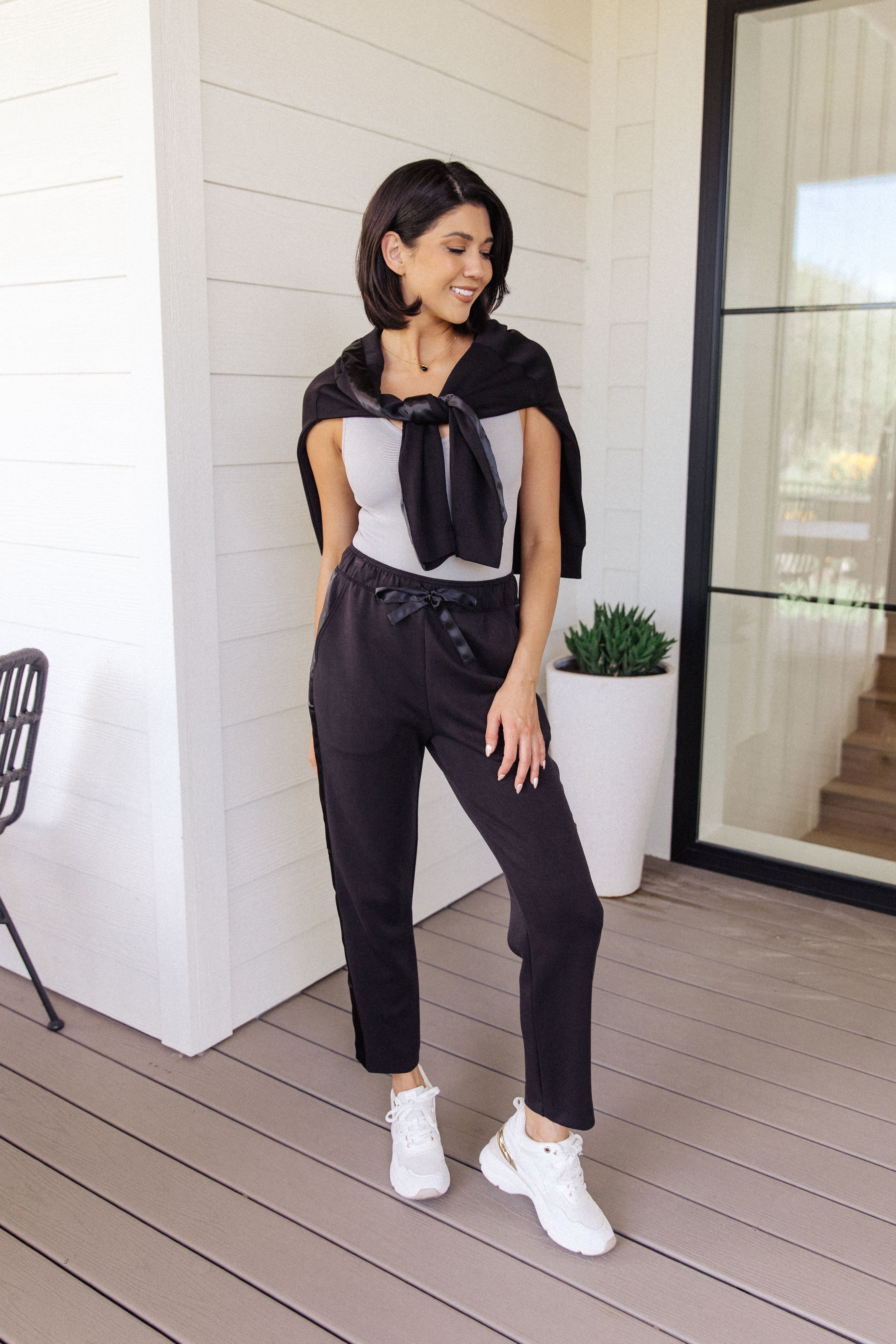 Woman with dark hair wearing a grey fitted tank top and black relaxed joggers standing on a porch smiling