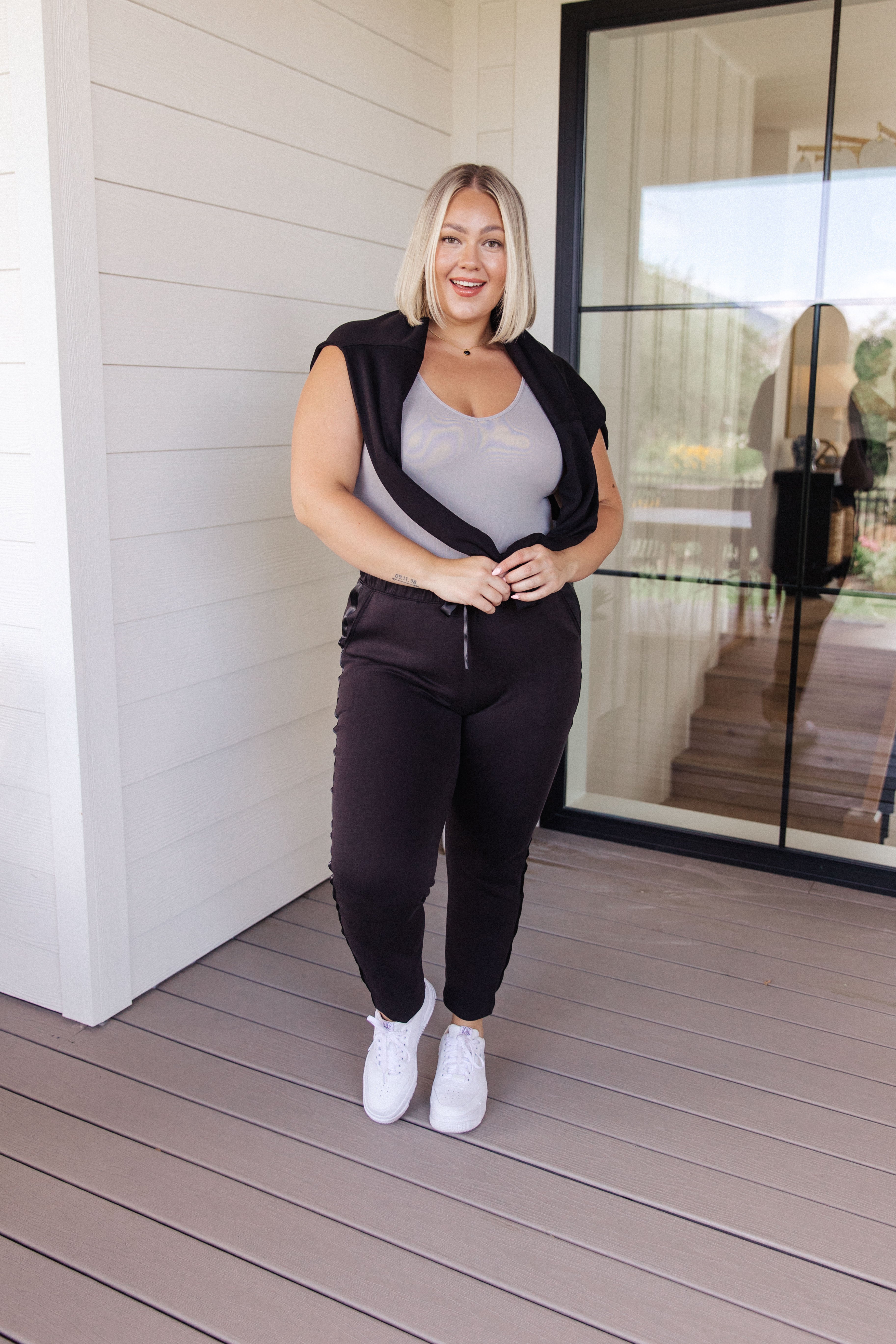 Smiling woman with blonde hair wearing a grey sleeveless top and black joggers standing on a wooden porch
