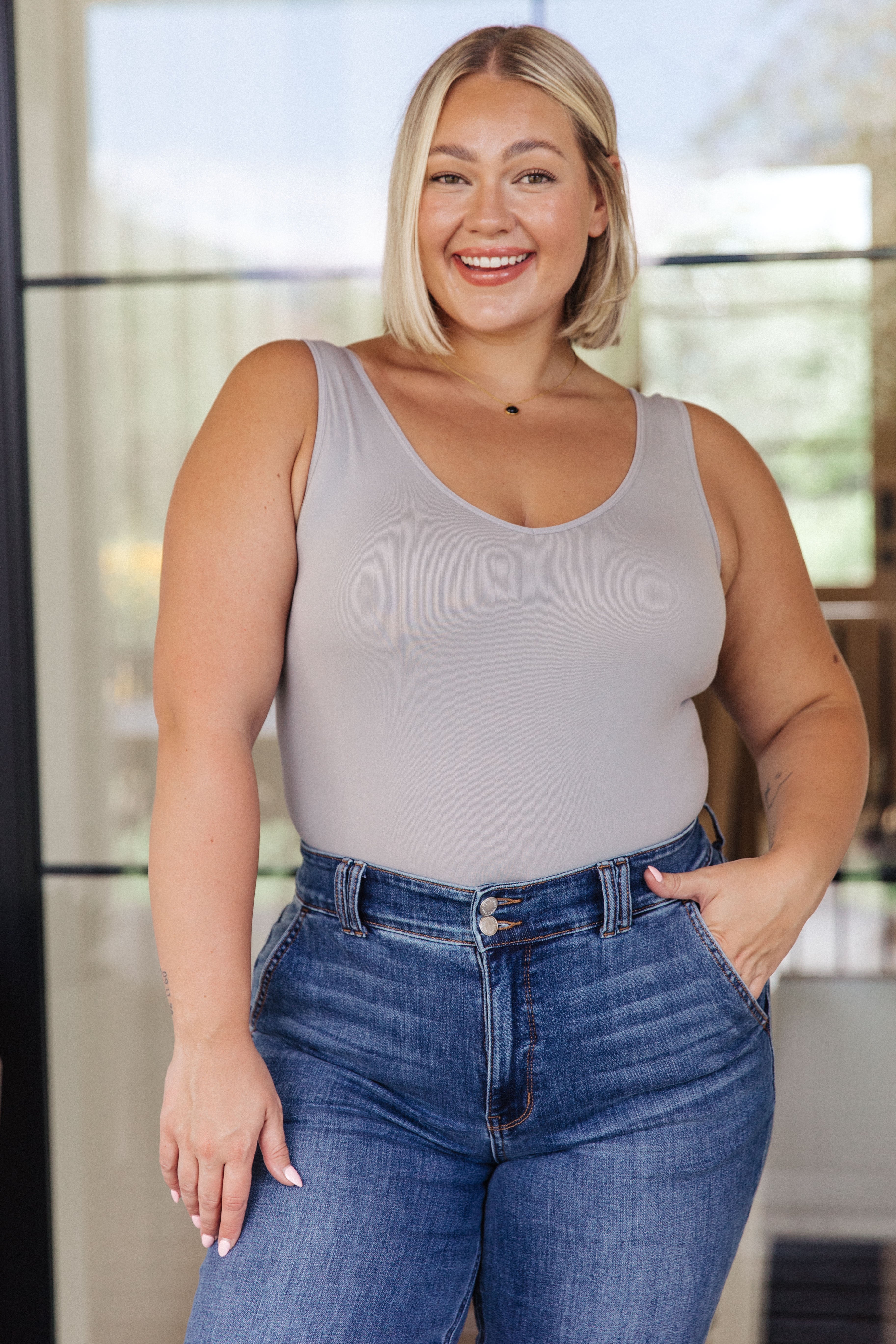 Smiling woman wearing a fitted grey sleeveless bodysuit tucked into high-waisted blue jeans with hands in pockets