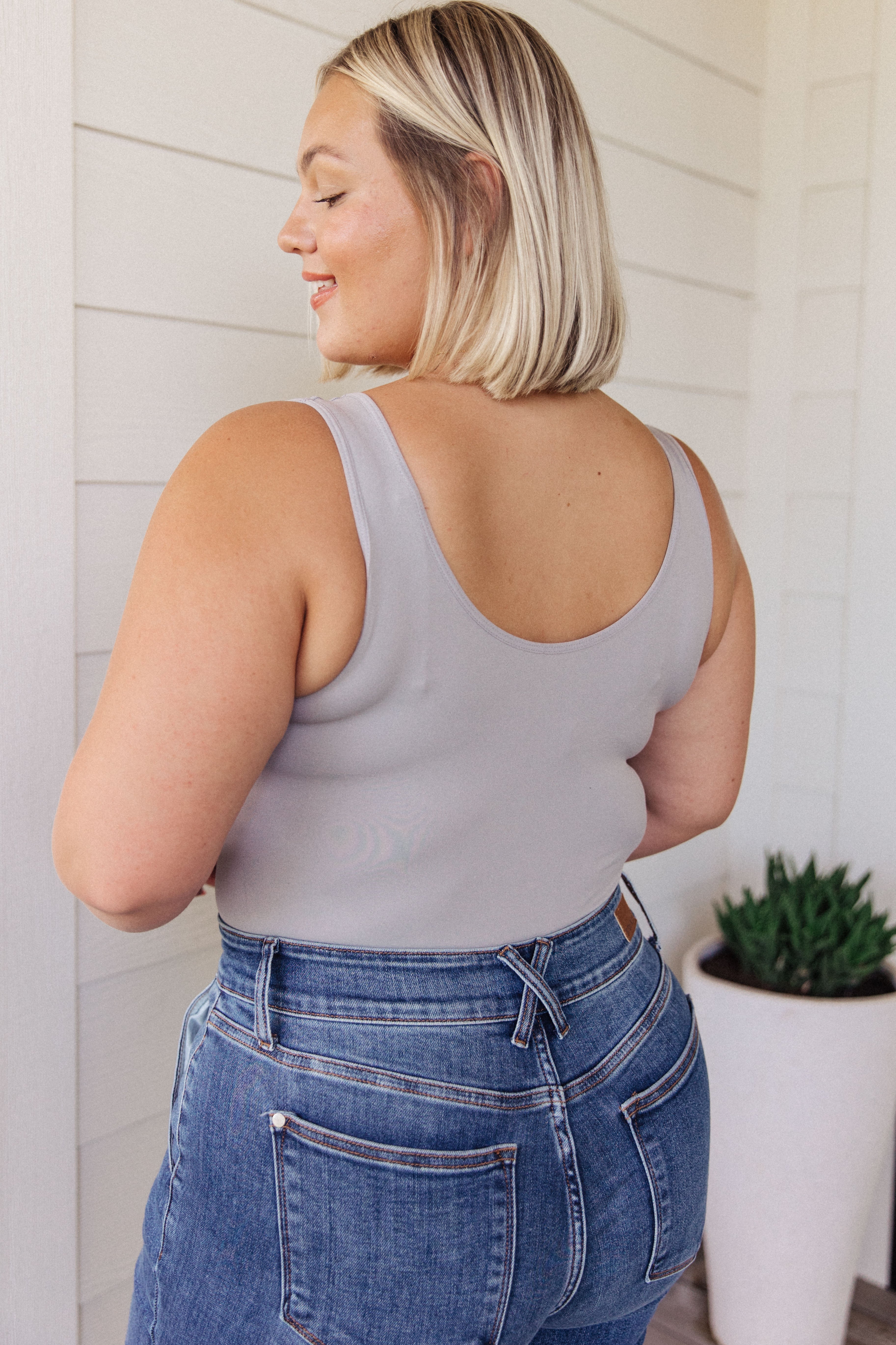 Plus size woman with blonde hair wearing a fitted grey sleeveless bodysuit and high-waisted blue jeans seen from the back
