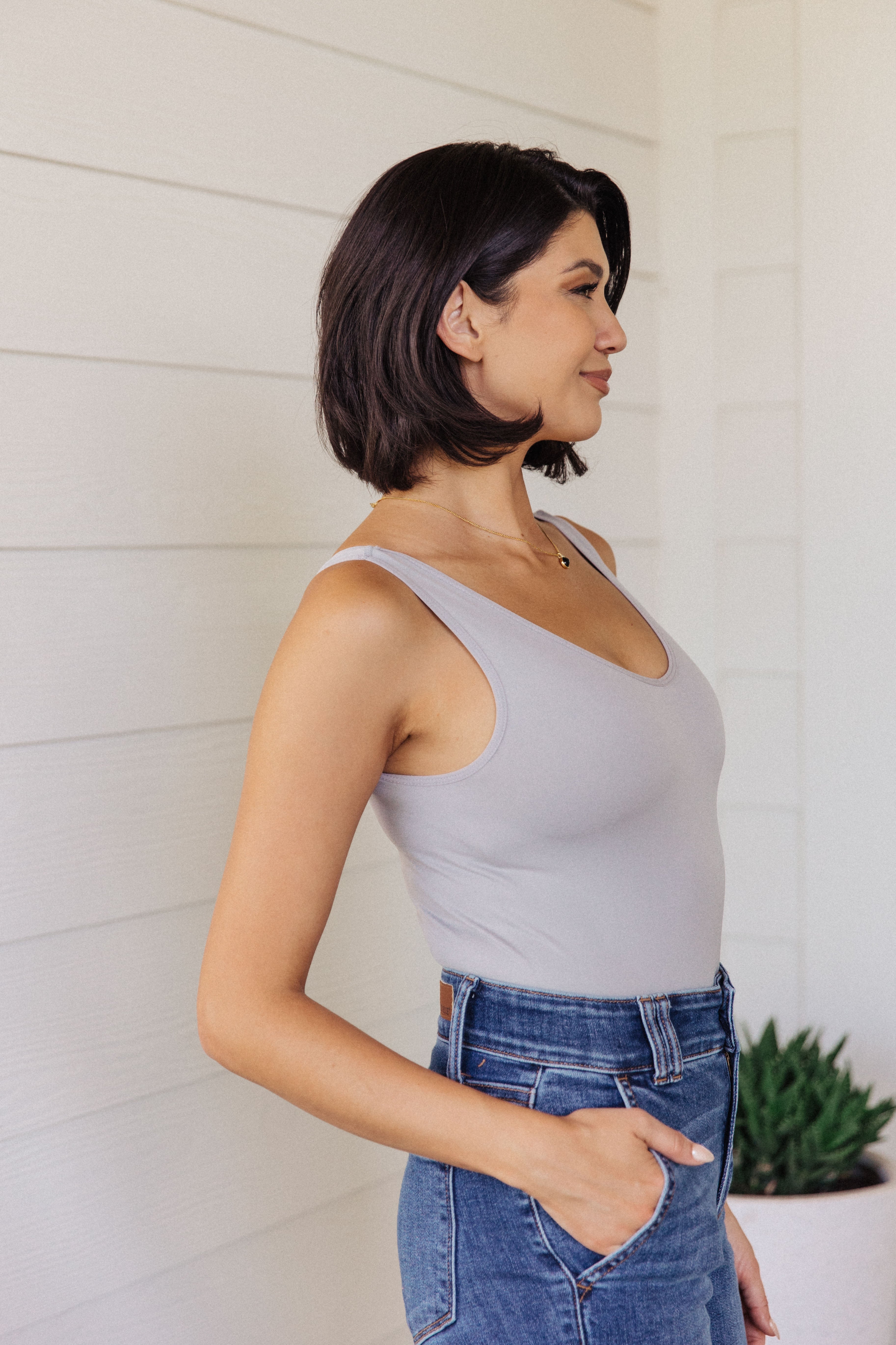 Woman wearing a fitted grey sleeveless bodysuit paired with high-waisted blue jeans standing sideways with one hand in pocket