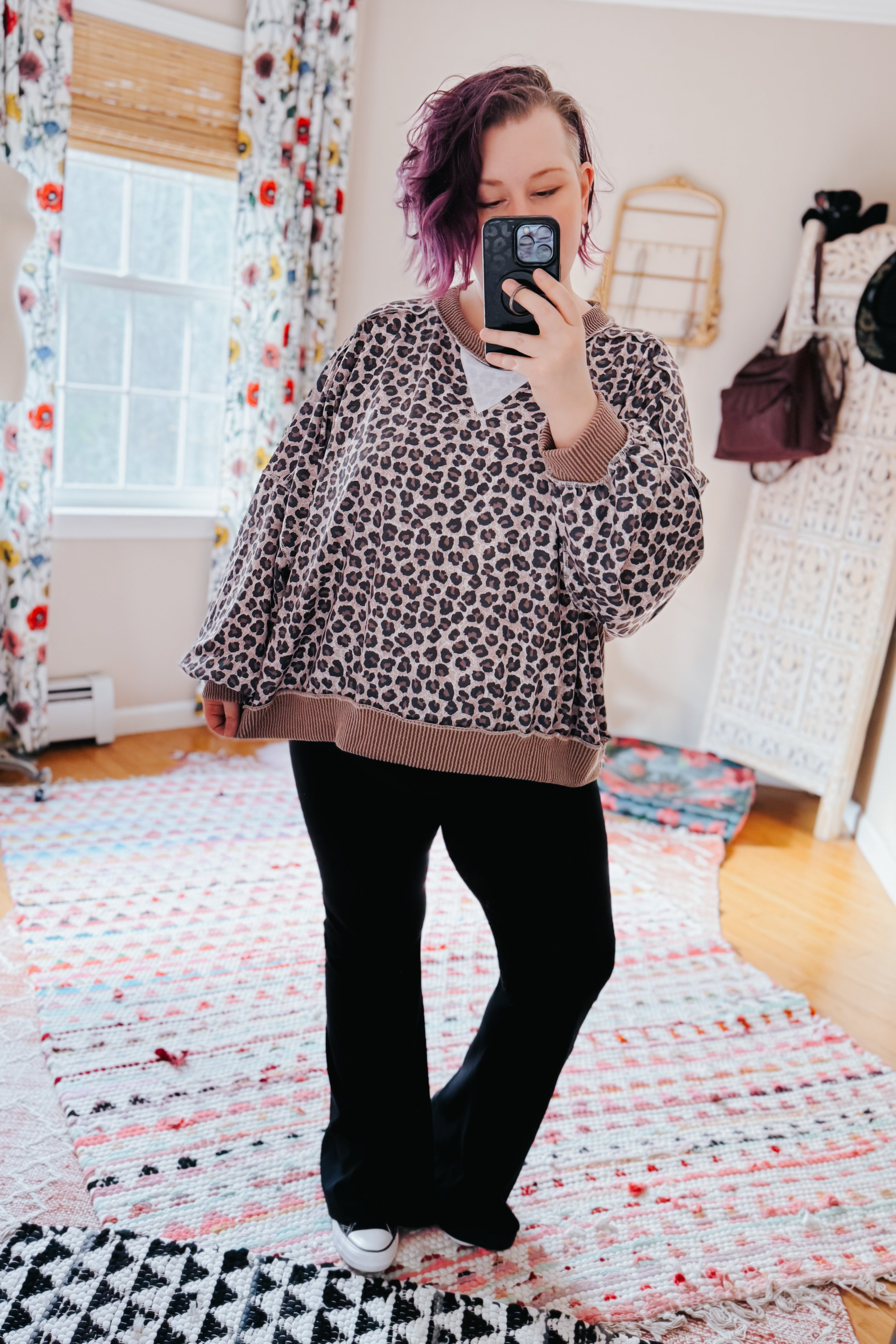 Woman with short purple hair wearing a brown leopard print oversized pullover and black pants taking a mirror selfie indoors