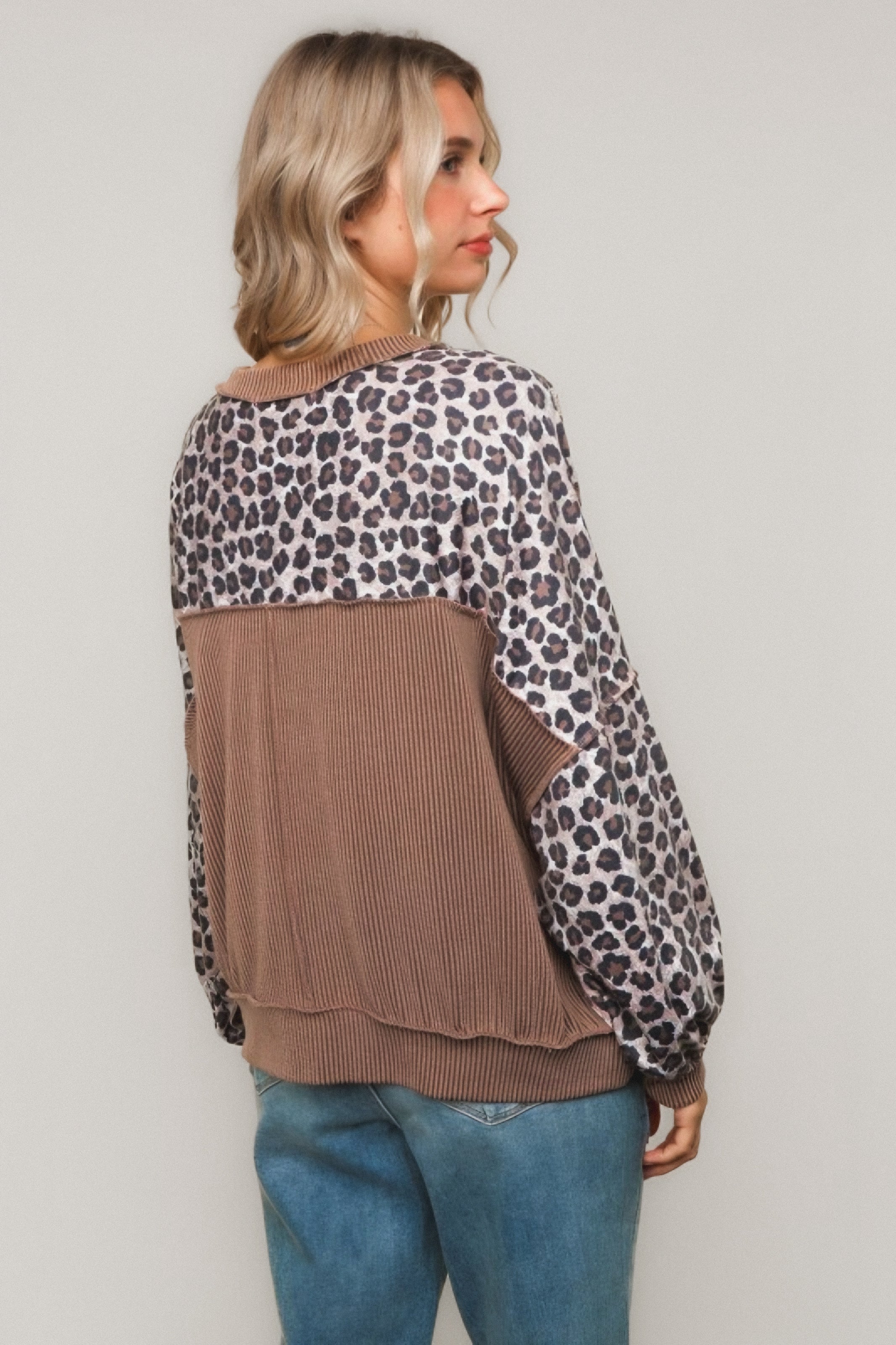 Woman with blonde hair wearing a brown pullover with leopard print yoke and sleeves paired with blue jeans standing against a wall