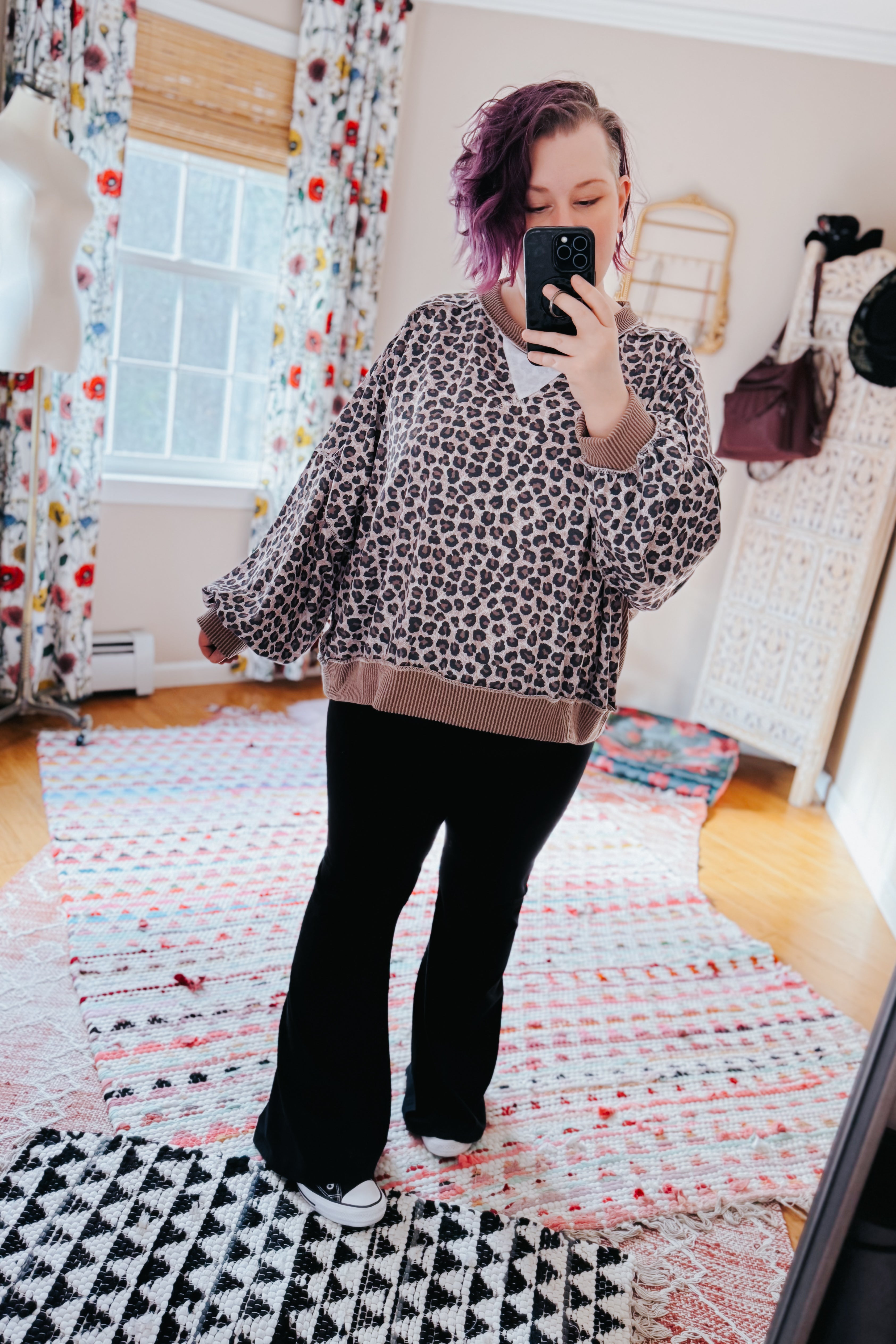 Woman with short purple hair wearing a brown leopard print pullover and black pants taking a mirror selfie indoors