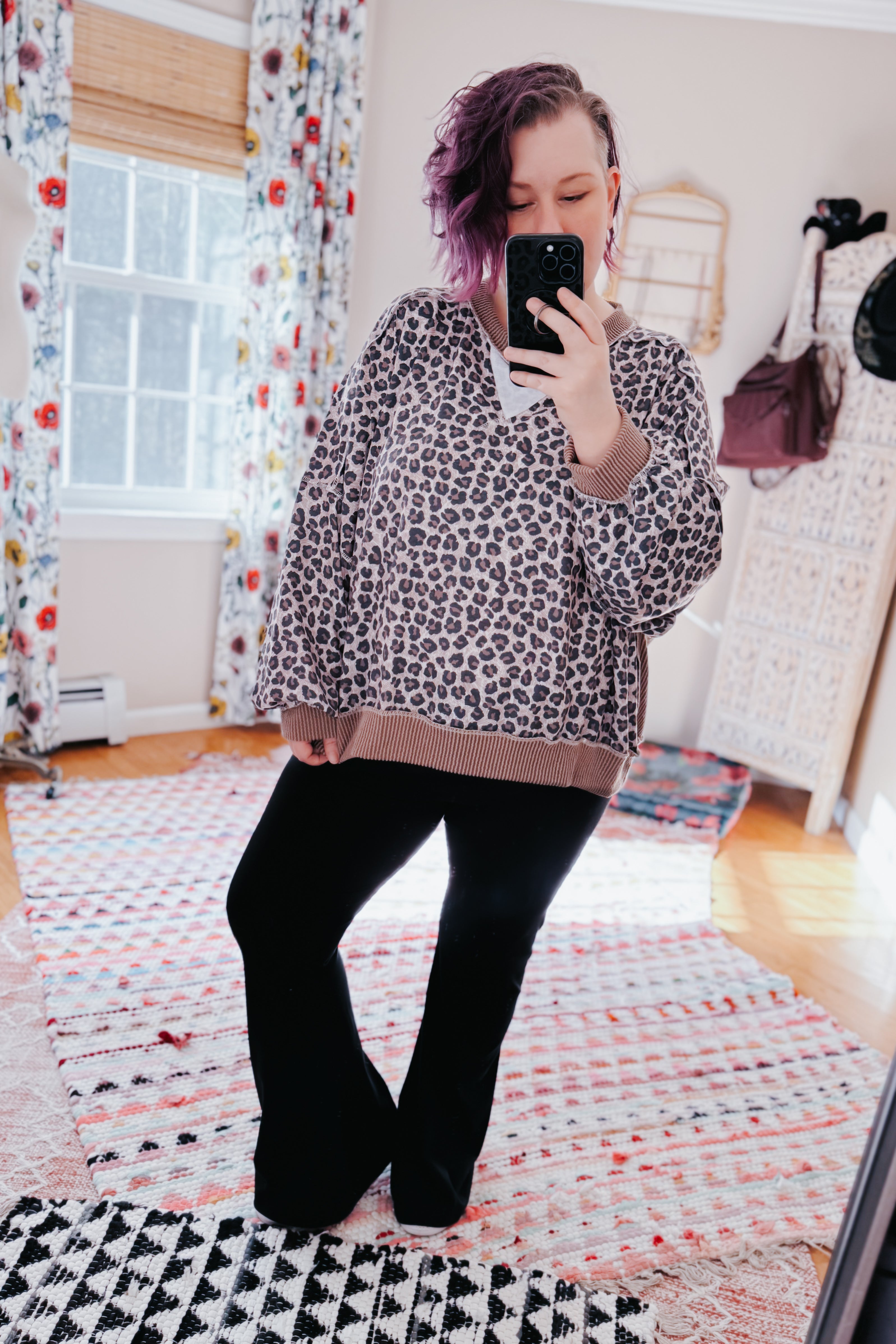 Woman with short purple hair wearing a brown leopard print pullover and black pants taking a mirror selfie indoors