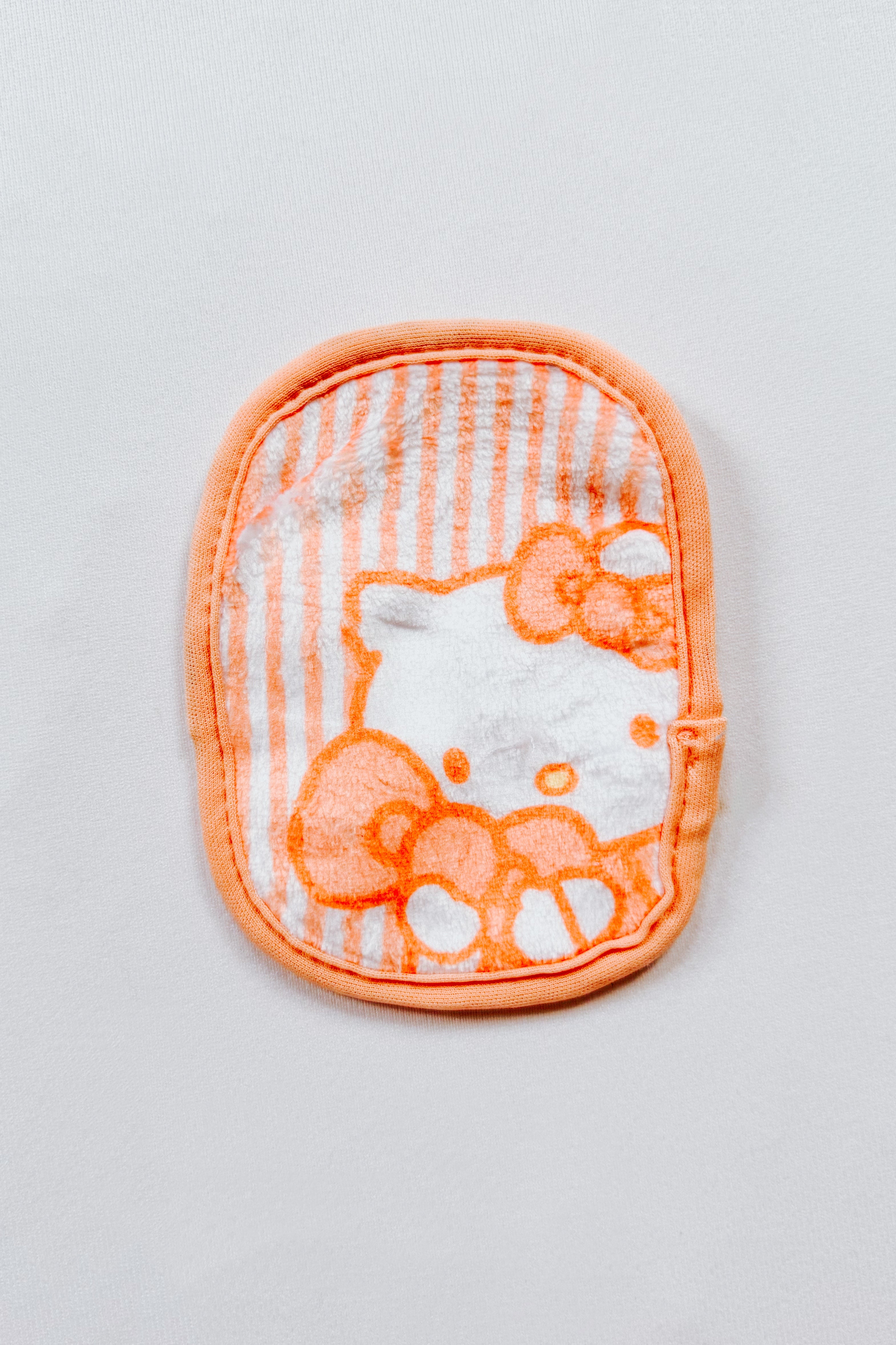 Orange and white striped makeup remover cloth featuring a partial Hello Kitty face with bow detail on white background