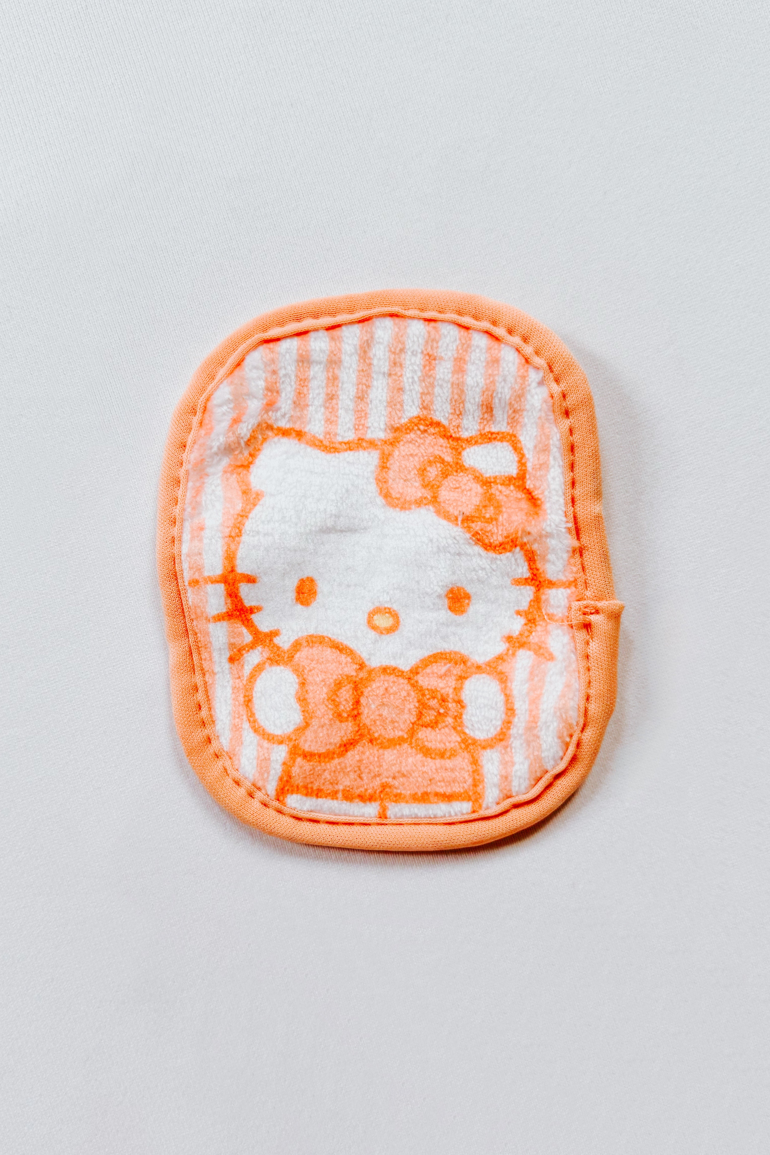 Small orange and white makeup eraser pad featuring a Hello Kitty face with striped background on white surface