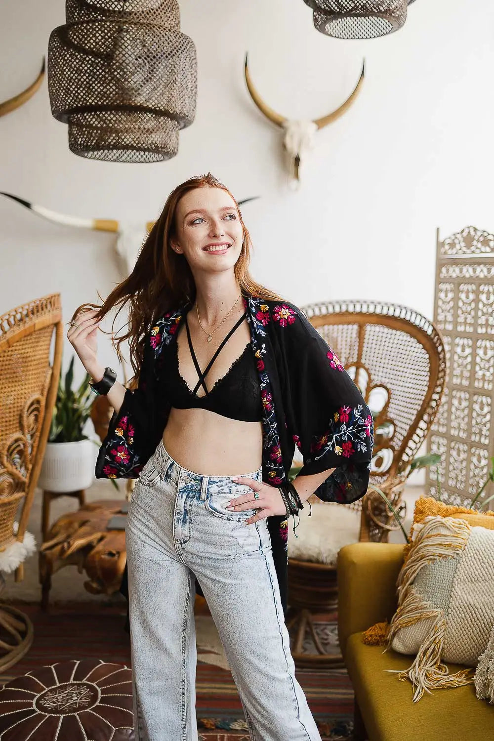 Smiling woman wearing a black embroidered duster with light wash jeans in a relaxed, eclectic indoor setting