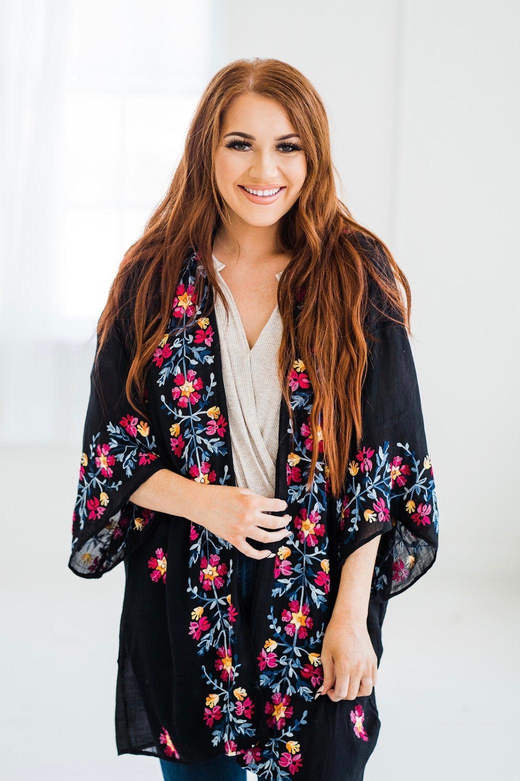 Woman wearing a black embroidered duster with colorful floral designs styled in a relaxed, eclectic look indoors