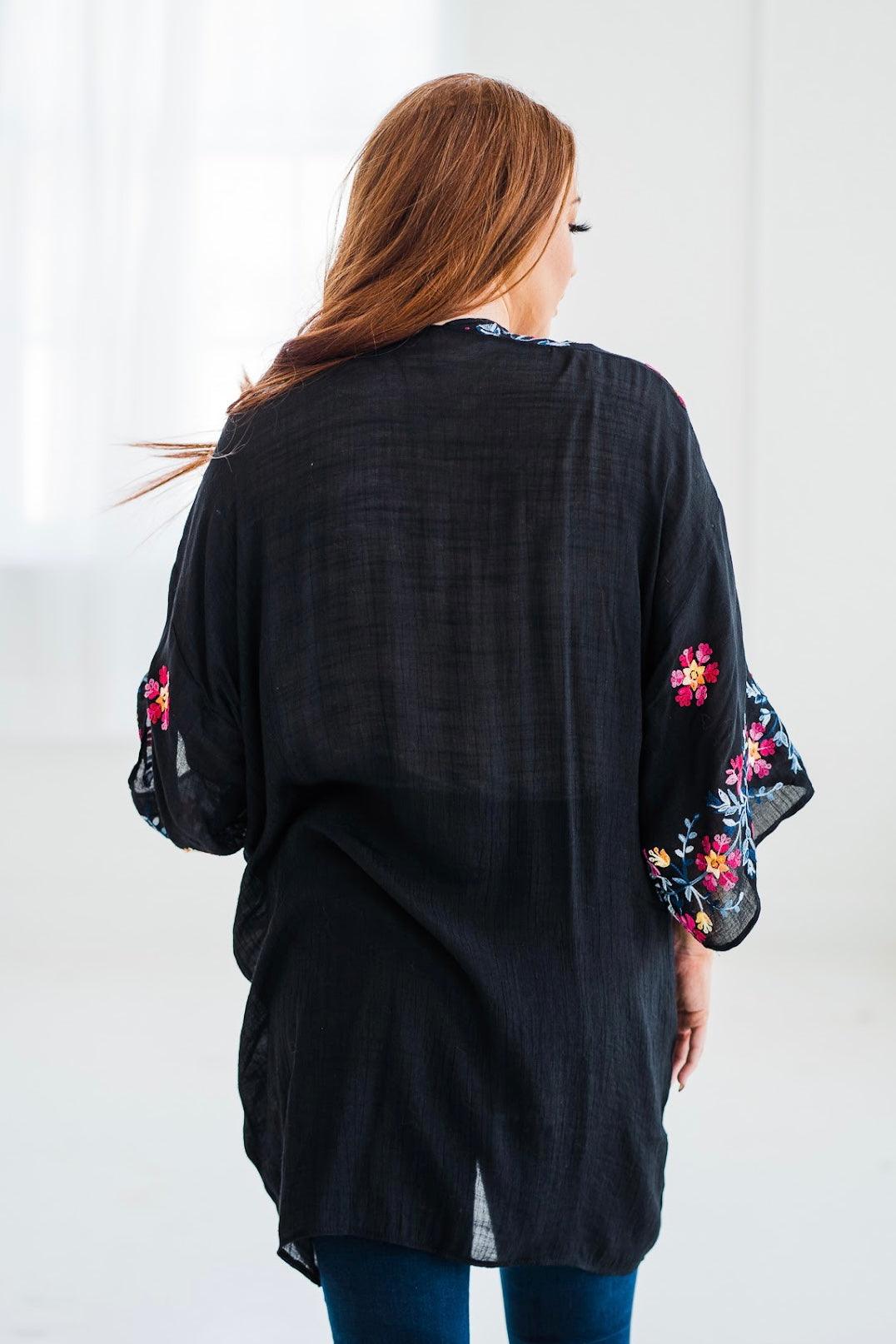 Back view of a black embroidered duster with colorful floral details on sleeves worn by a woman indoors