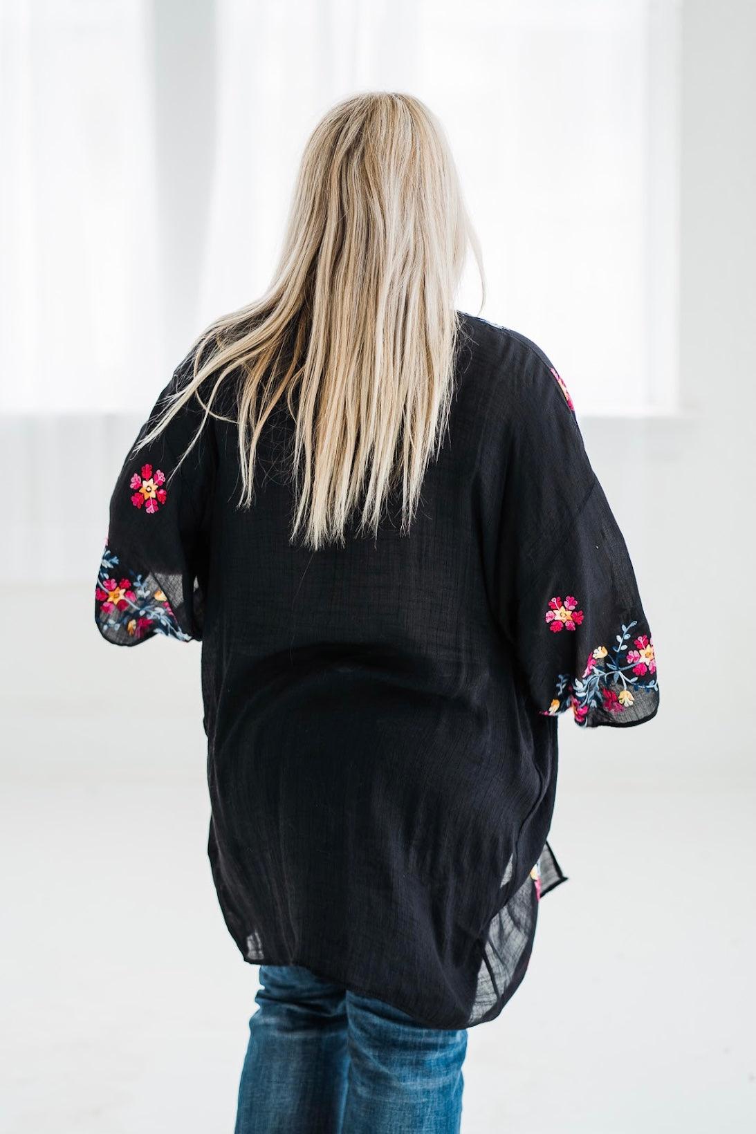 Back view of a woman wearing a black embroidered duster with colorful floral details and blue jeans indoors
