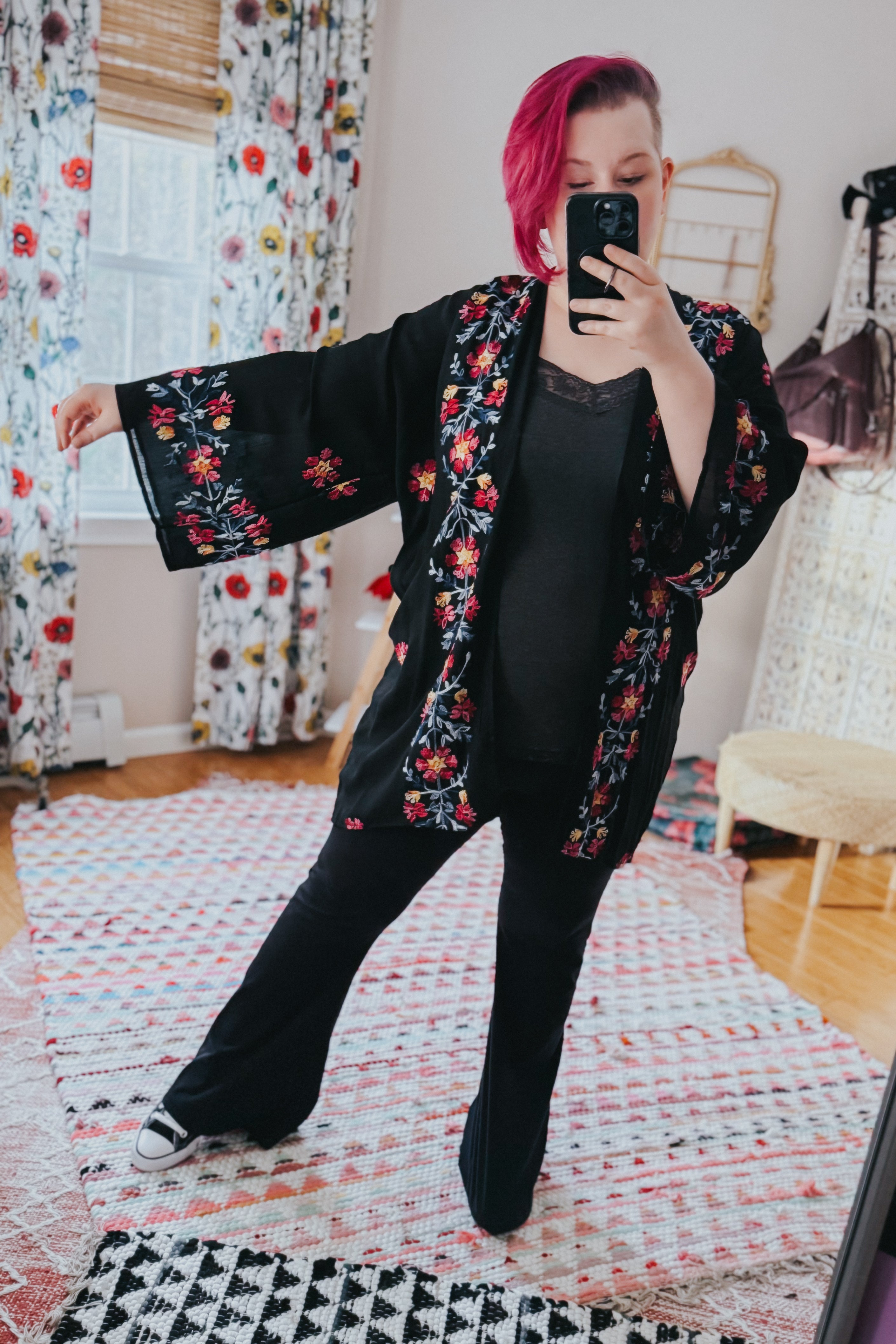 Woman with pink hair wearing a black embroidered duster and black pants taking a mirror selfie indoors