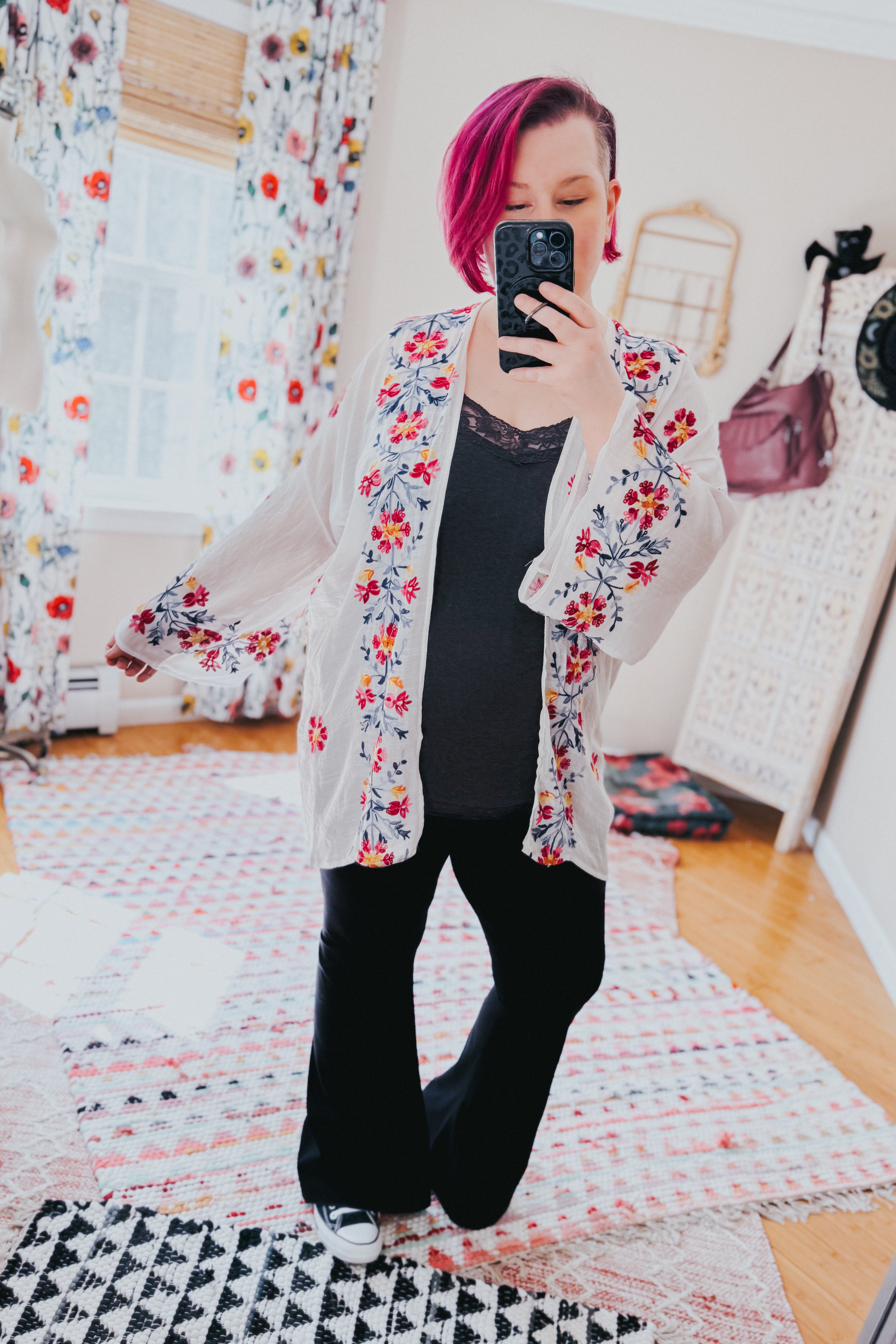 Woman with magenta hair wearing an ivory embroidered duster over a black top and pants, taking a mirror selfie indoors
