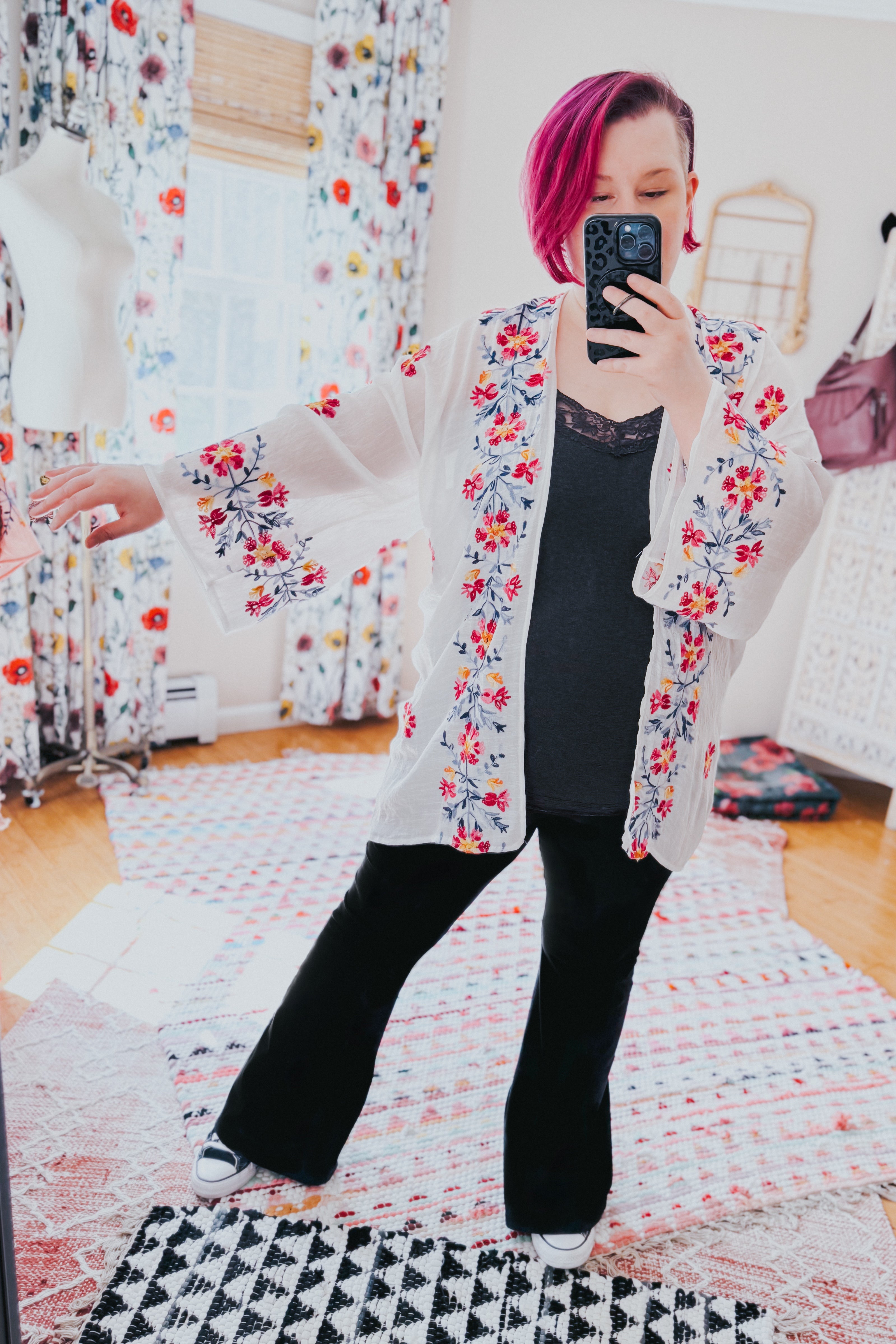 Woman wearing an ivory embroidered duster over black top and pants, posing in a bright room with patterned rugs and decor