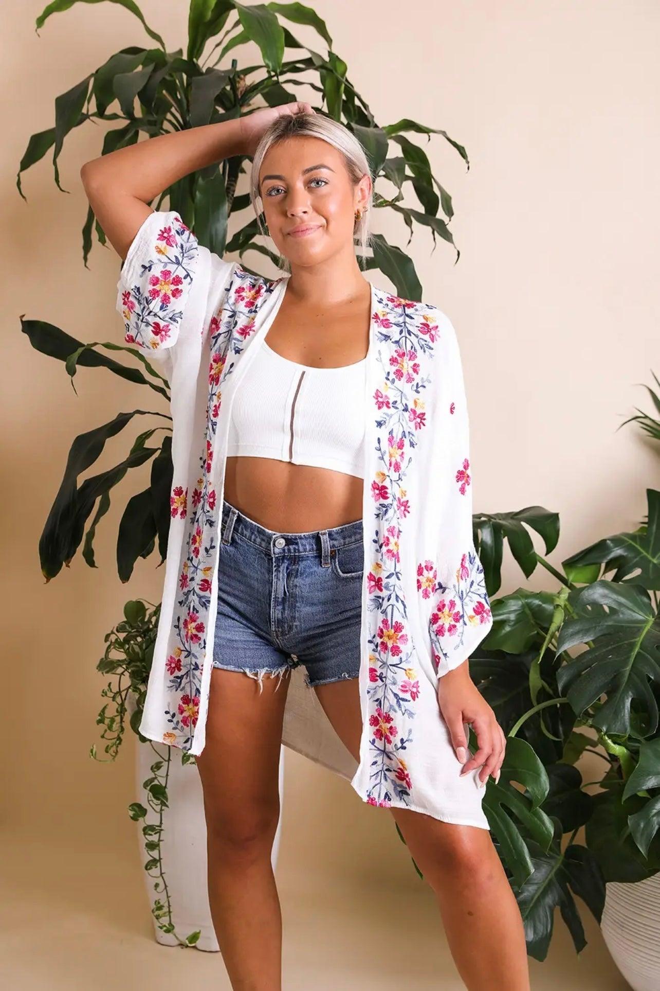 Woman wearing an ivory embroidered duster with floral designs over a white crop top and denim shorts standing indoors
