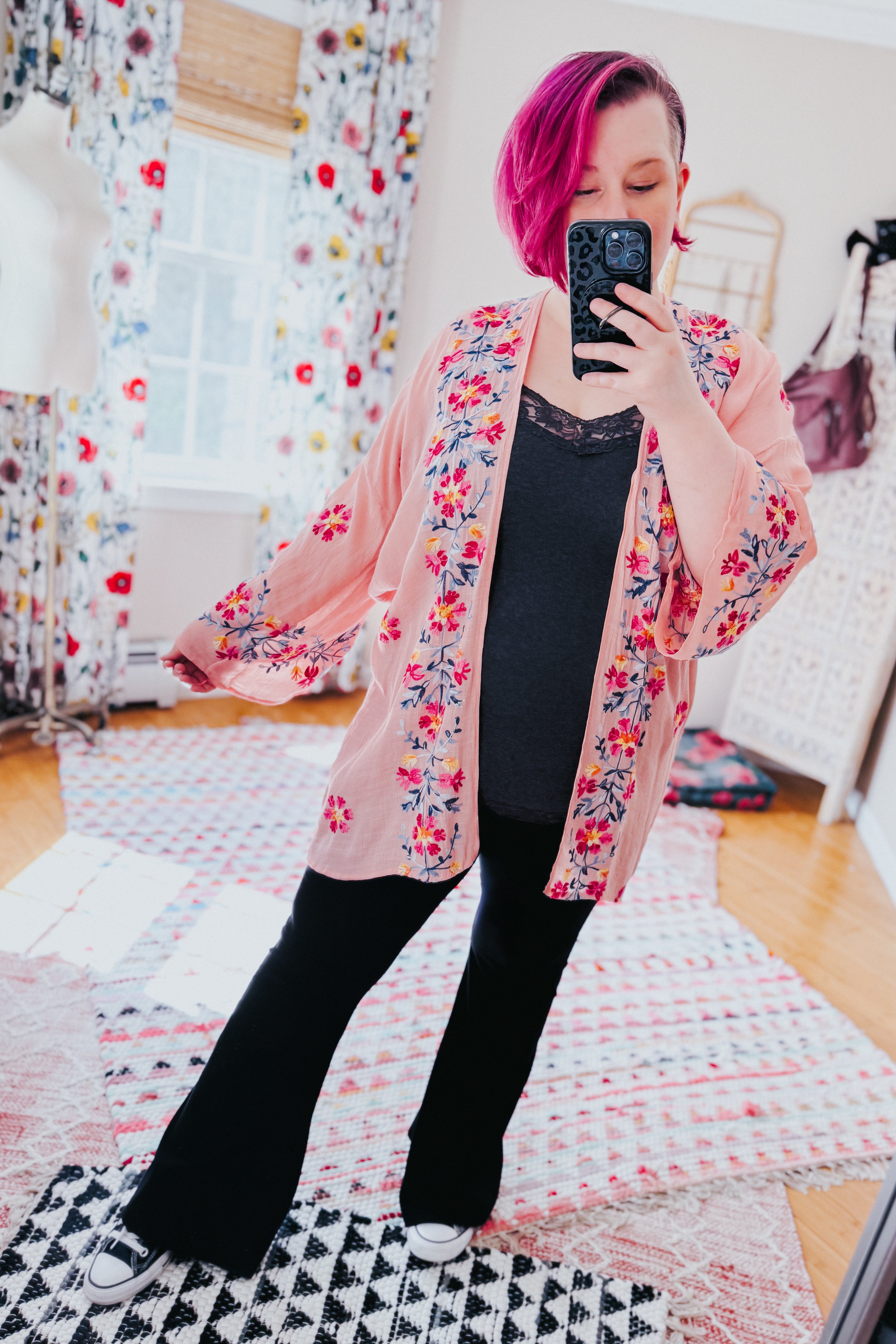 Woman with magenta hair wearing a rose embroidered duster with floral patterns over black top and black pants indoors