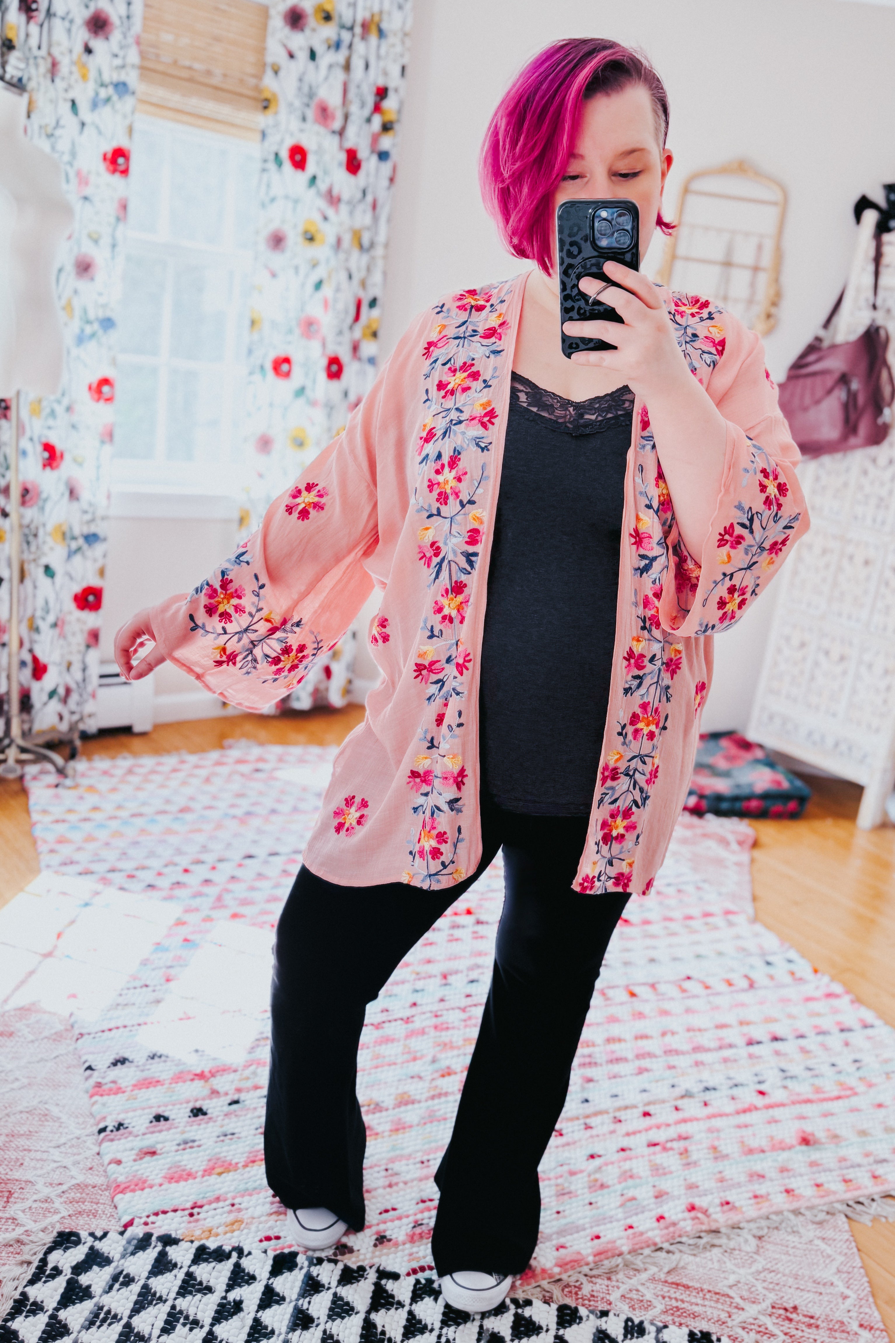 Woman with magenta hair wearing a rose embroidered duster with floral patterns over a black top and pants in a bright room