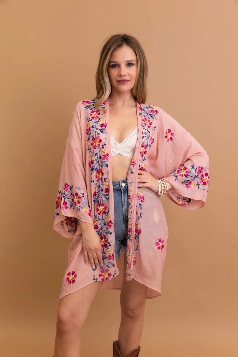Woman wearing a rose embroidered duster with floral patterns over a white lace bralette and denim shorts