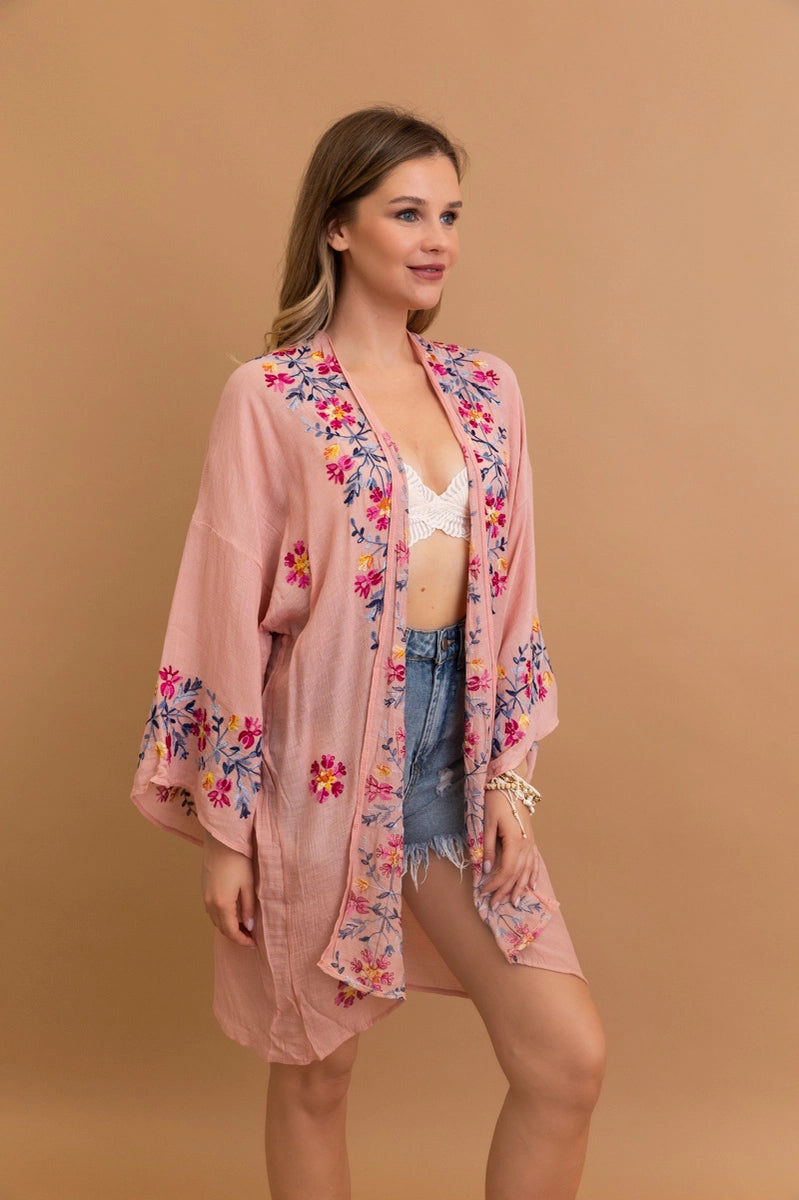 Woman wearing a rose embroidered duster with floral patterns over a white lace top and denim shorts against tan background