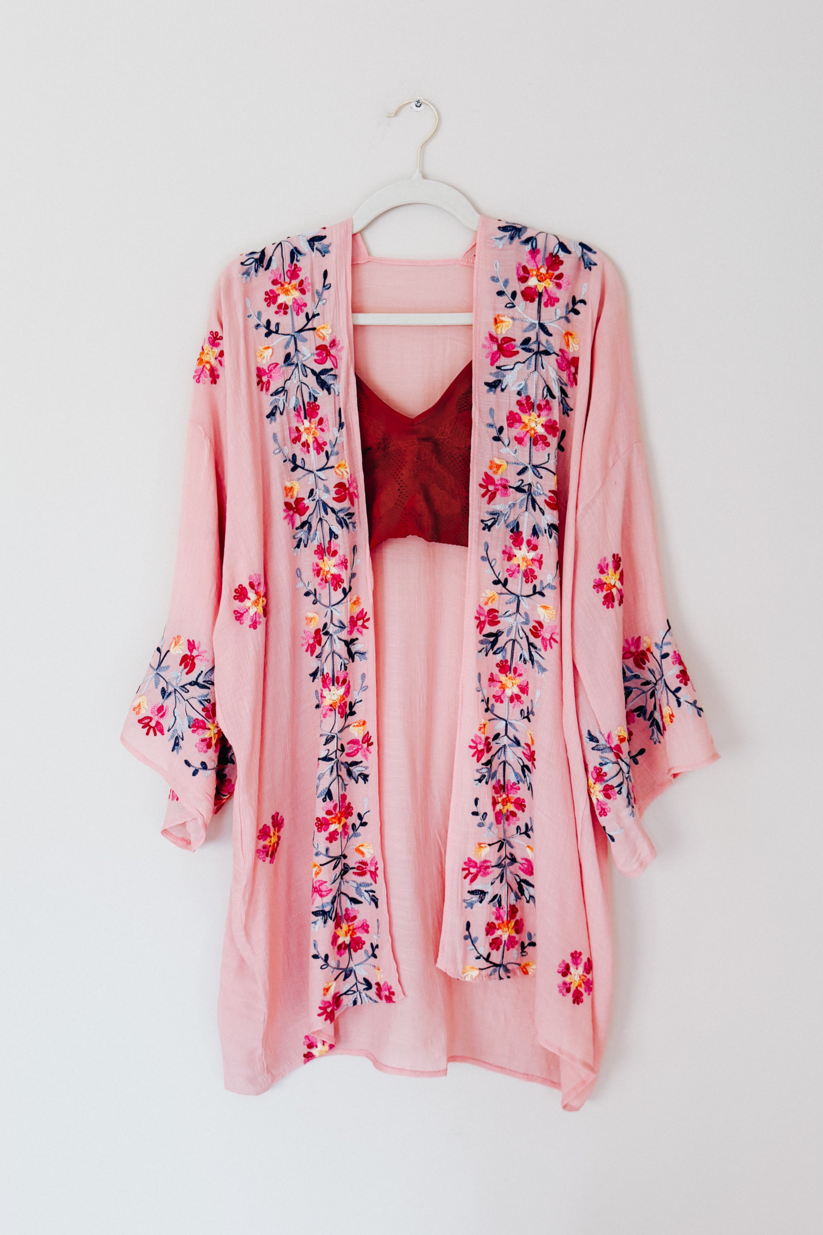 Rose-colored embroidered duster with floral patterns along front and sleeves hanging on a white hanger against plain wall