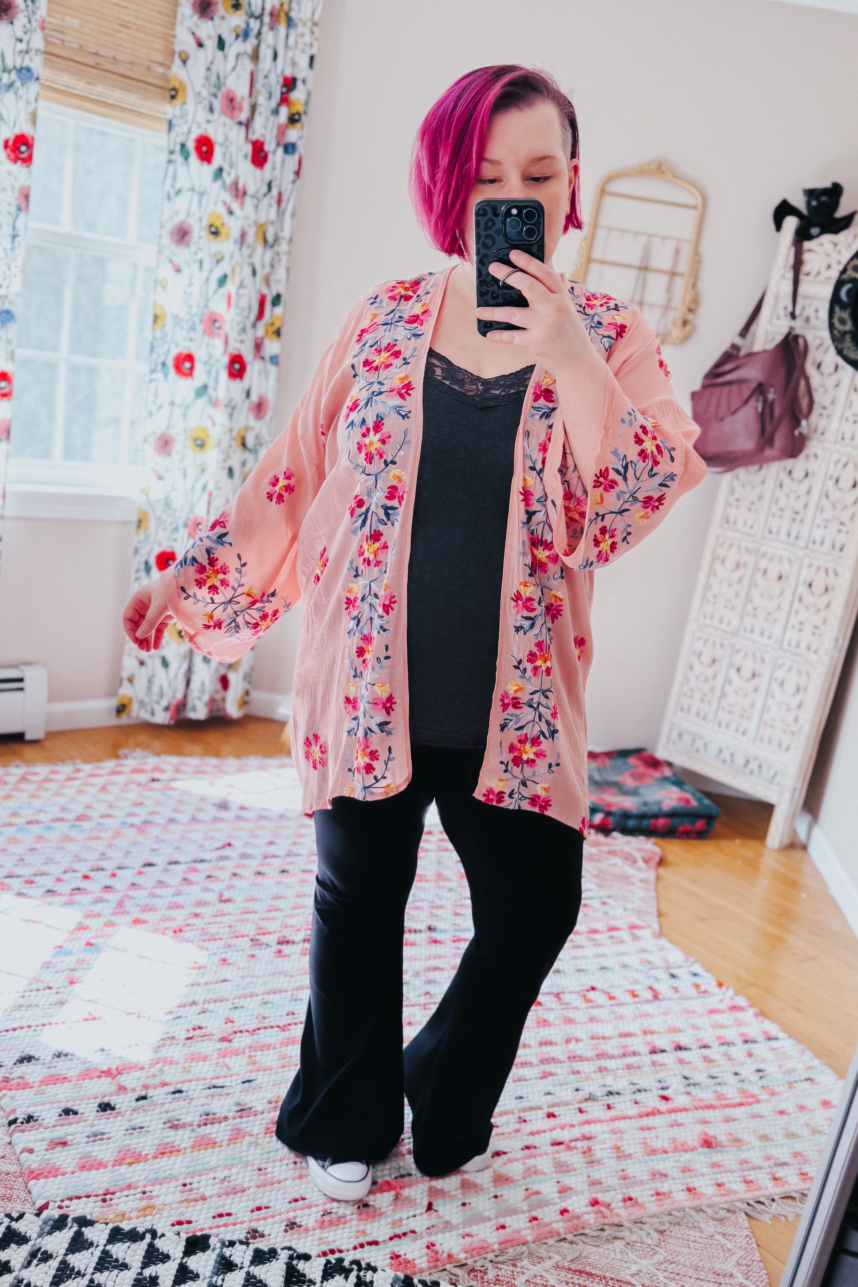 Woman with magenta hair wearing a rose embroidered duster over black lace-trim top and flared pants in cozy room