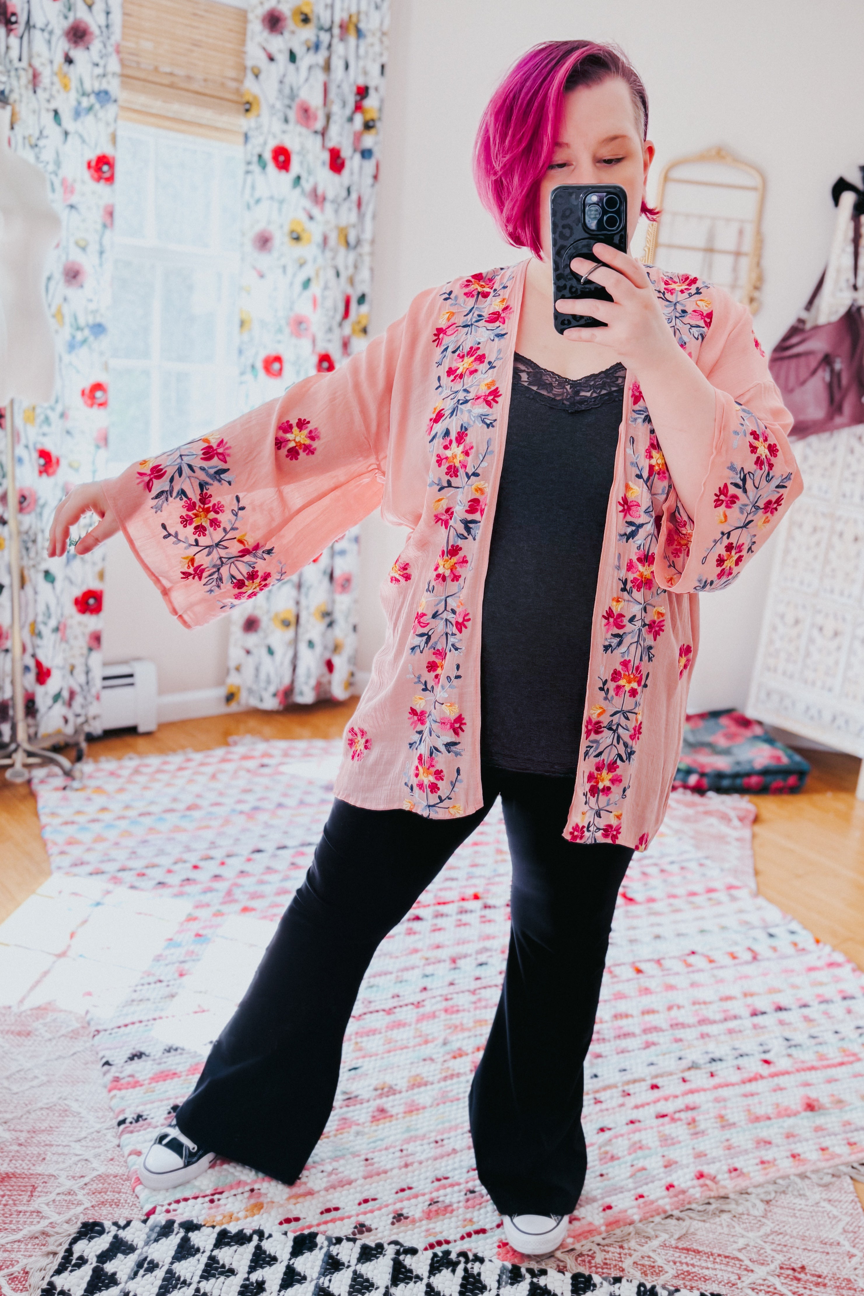 Woman with magenta hair wearing a rose embroidered duster over a black lace-trim top and black pants indoors