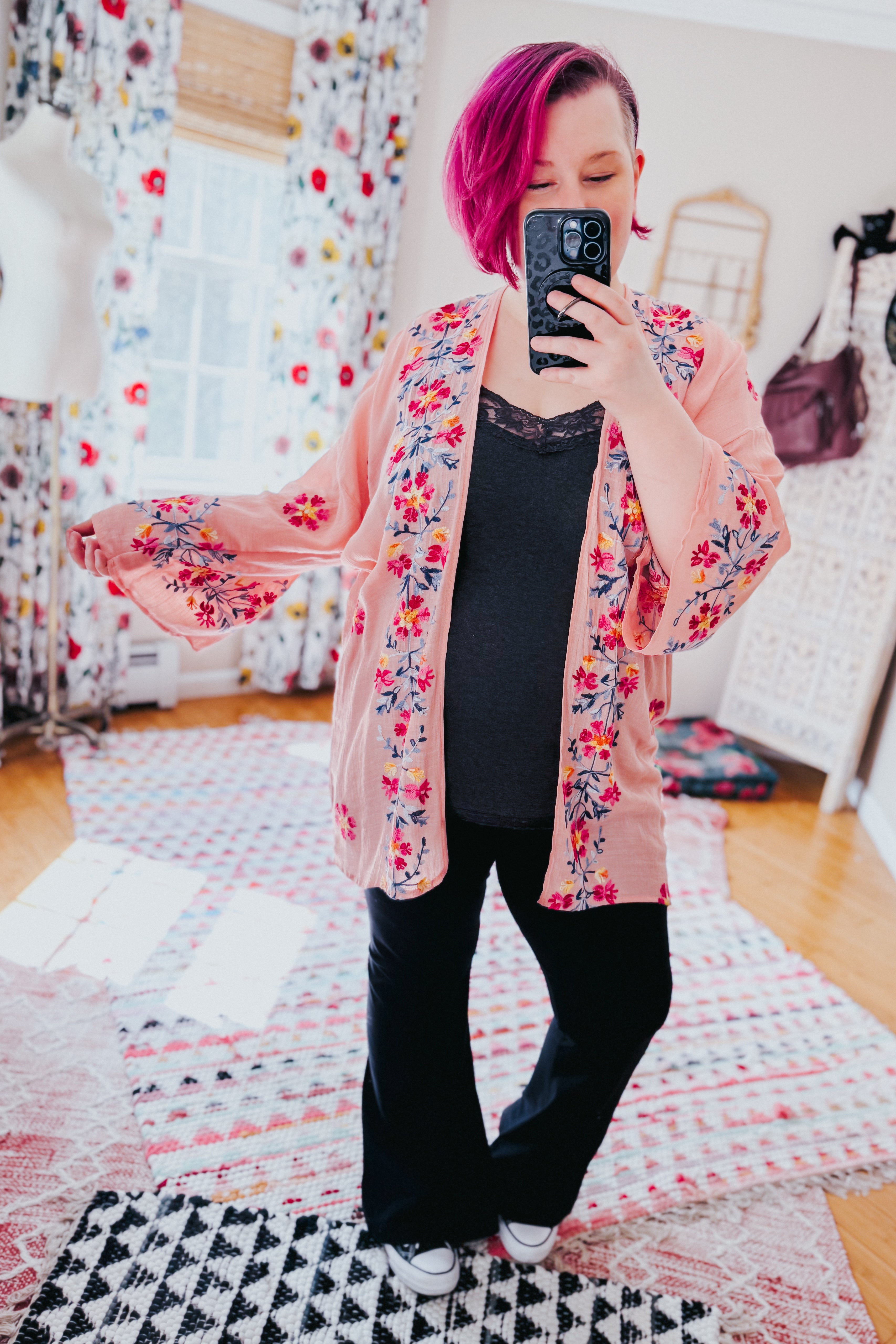 Woman with magenta hair wearing a rose embroidered duster over a black lace-trim top and pants, taking a mirror selfie indoors