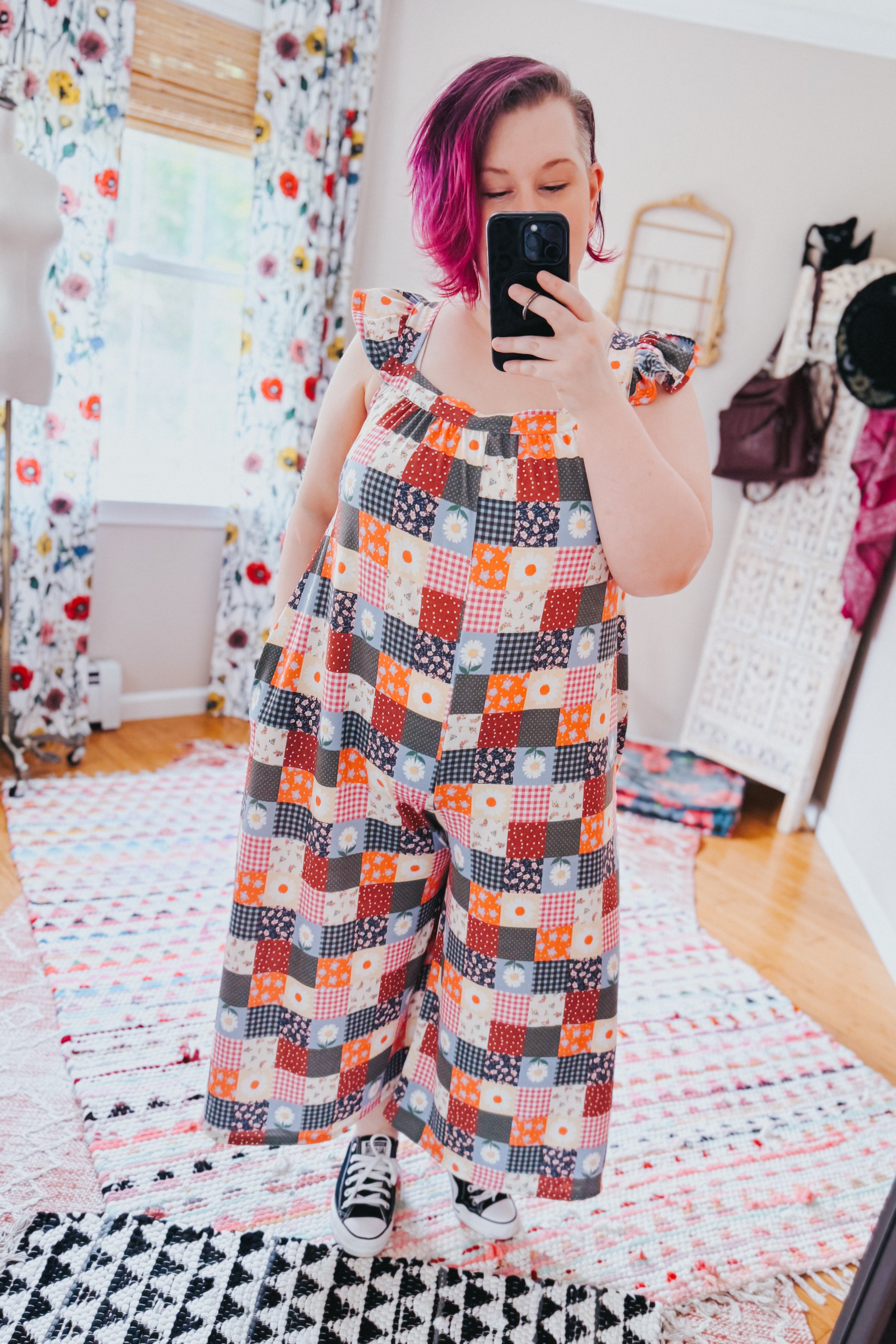Woman with magenta hair wearing a multicolor ruffle sleeve patchwork jumpsuit and black sneakers taking a mirror selfie indoors