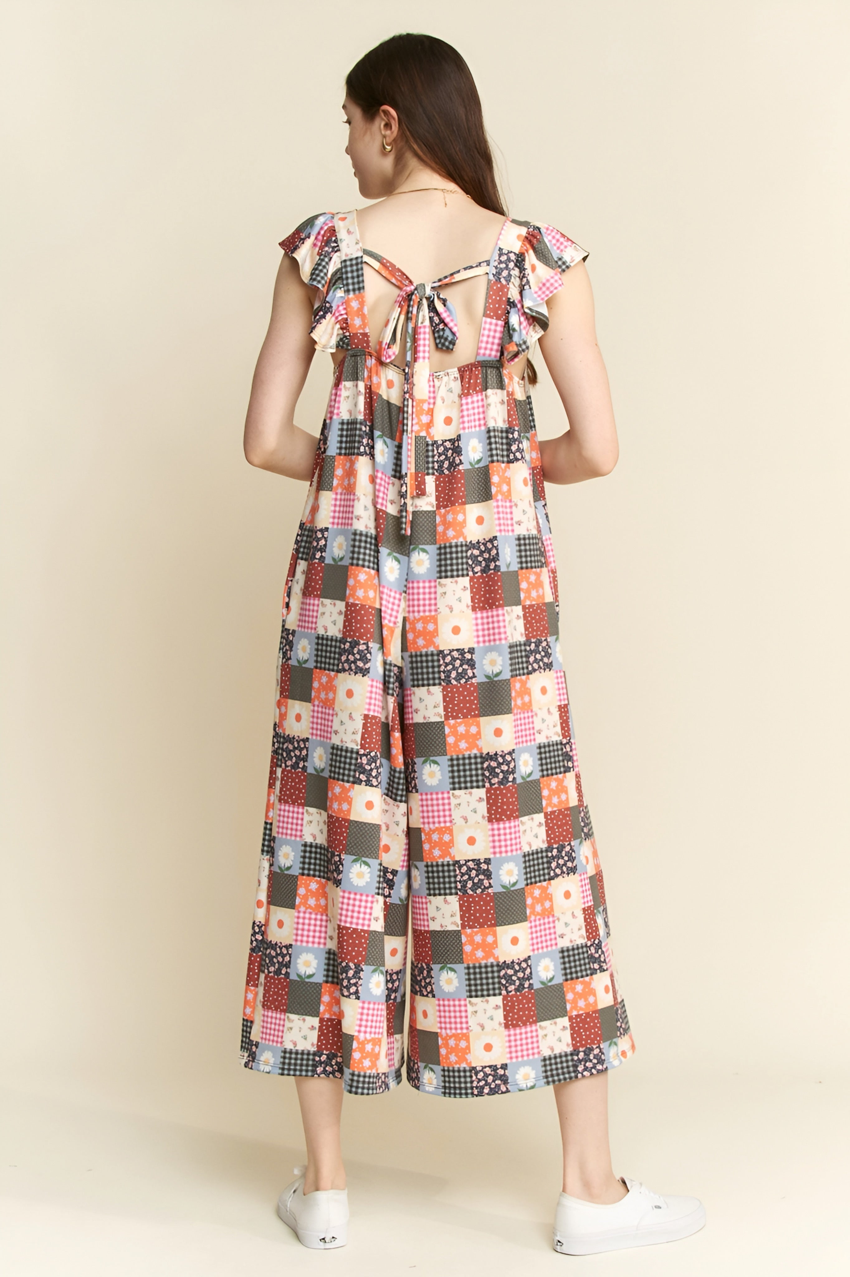 Woman wearing a multicolor patchwork jumpsuit with ruffle sleeves and an open back tied with a bow, standing indoors