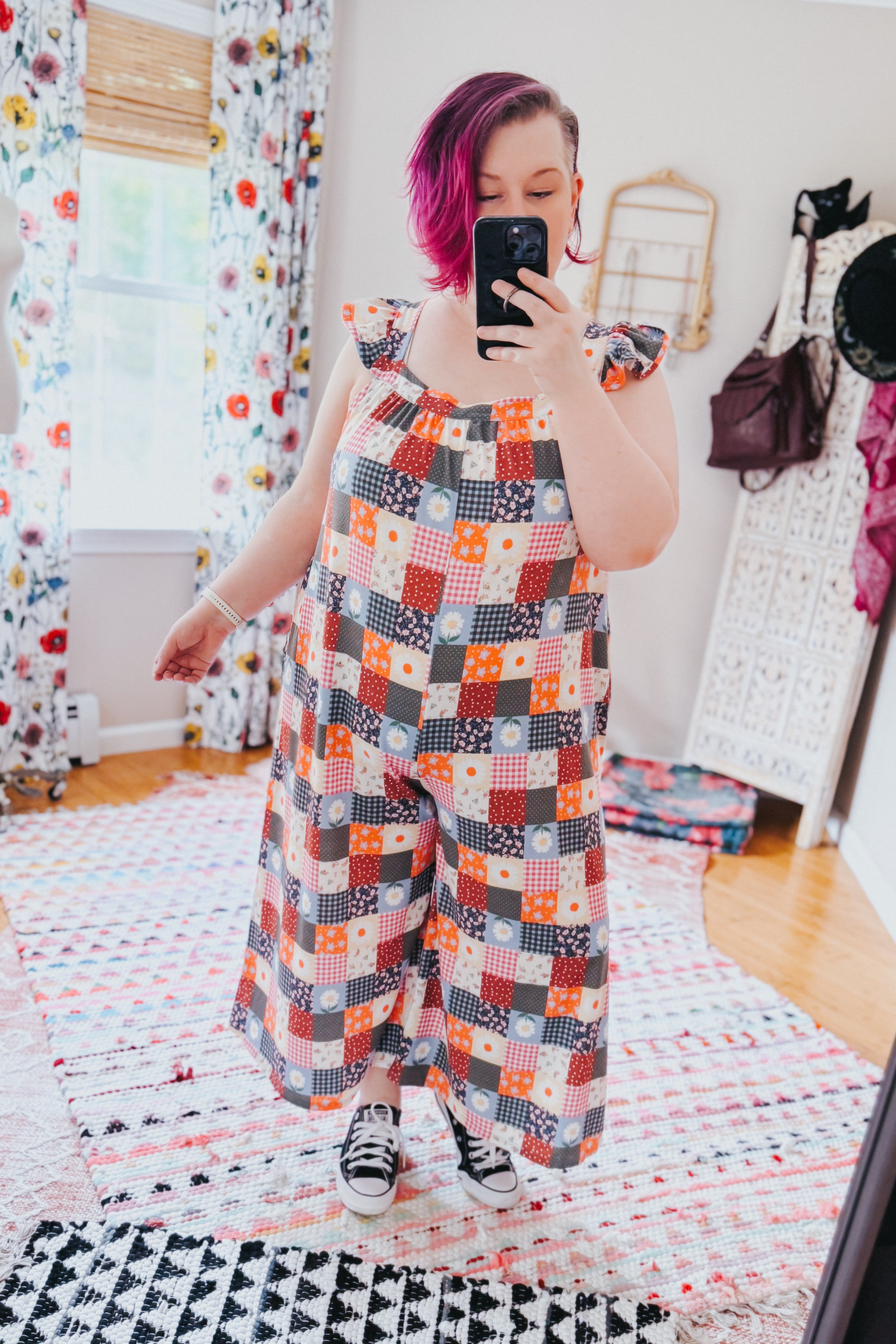 Woman with magenta hair wearing a colorful patchwork ruffle sleeve jumpsuit and black sneakers in bright room with floral curtains