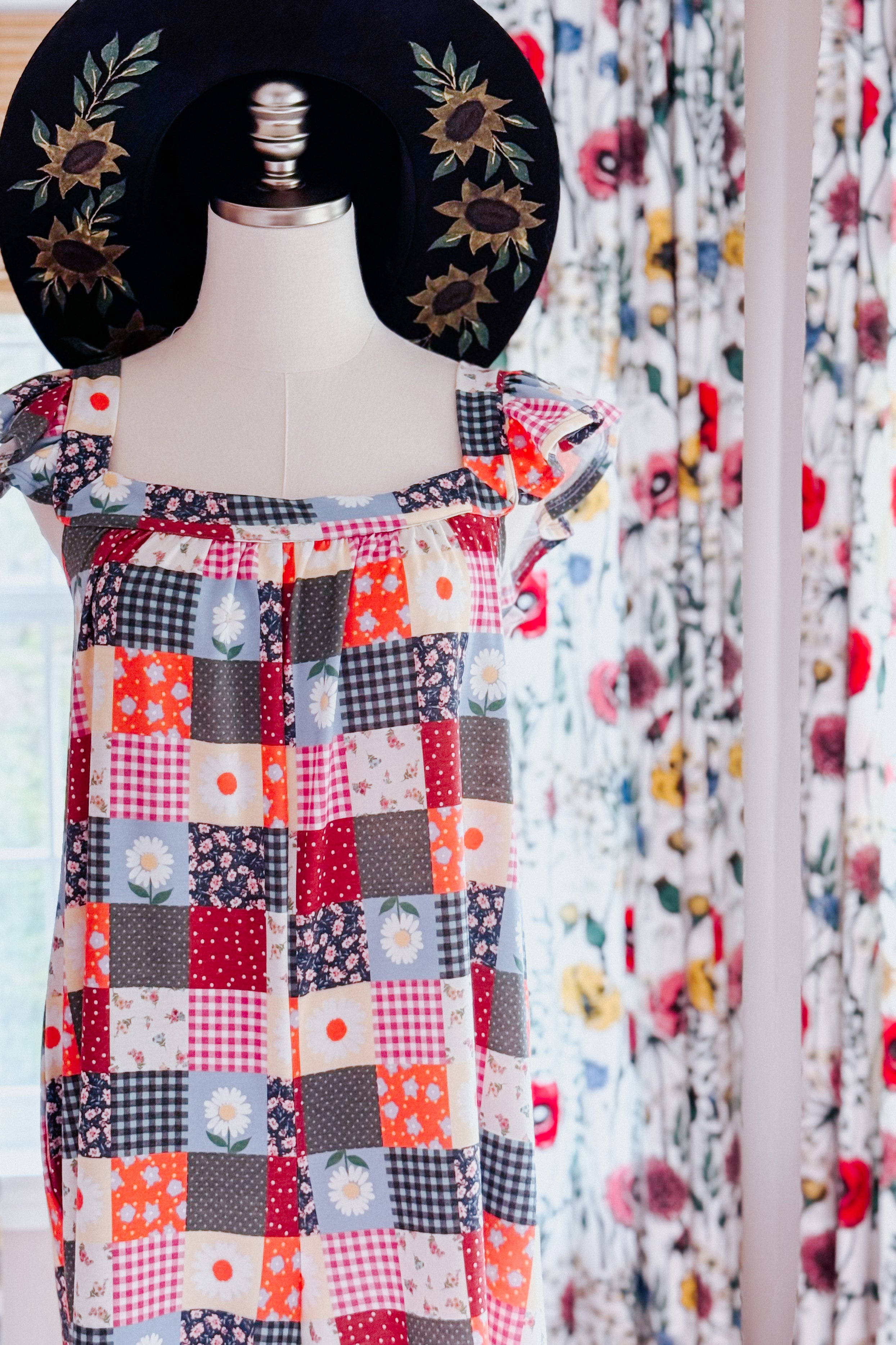 Mannequin wearing a colorful patchwork jumpsuit with ruffle sleeves and a black wide-brim hat embroidered with sunflowers