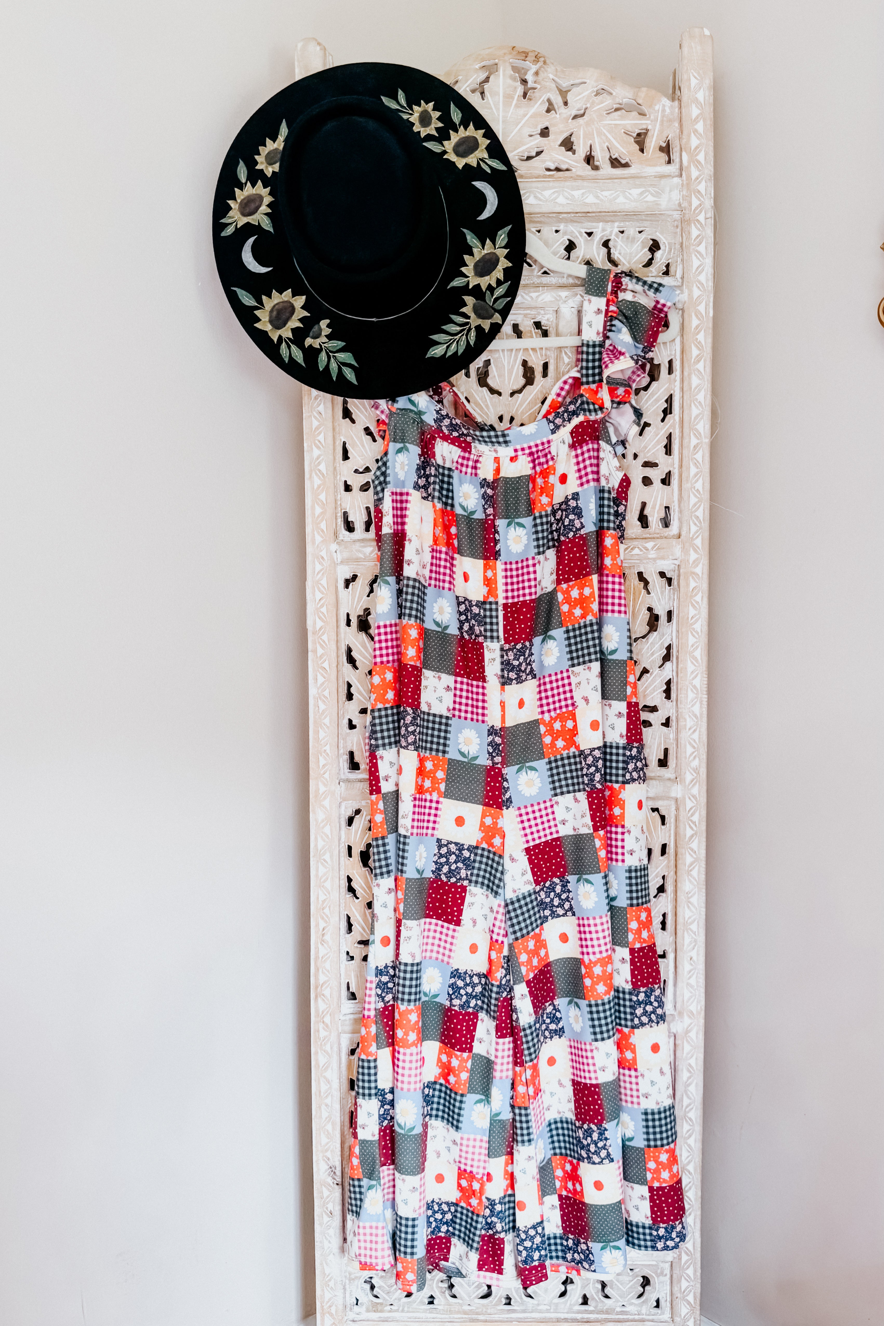 Multicolor patchwork jumpsuit with ruffle sleeves hanging on carved white wooden panel next to black hat with embroidered sunflowers and moons