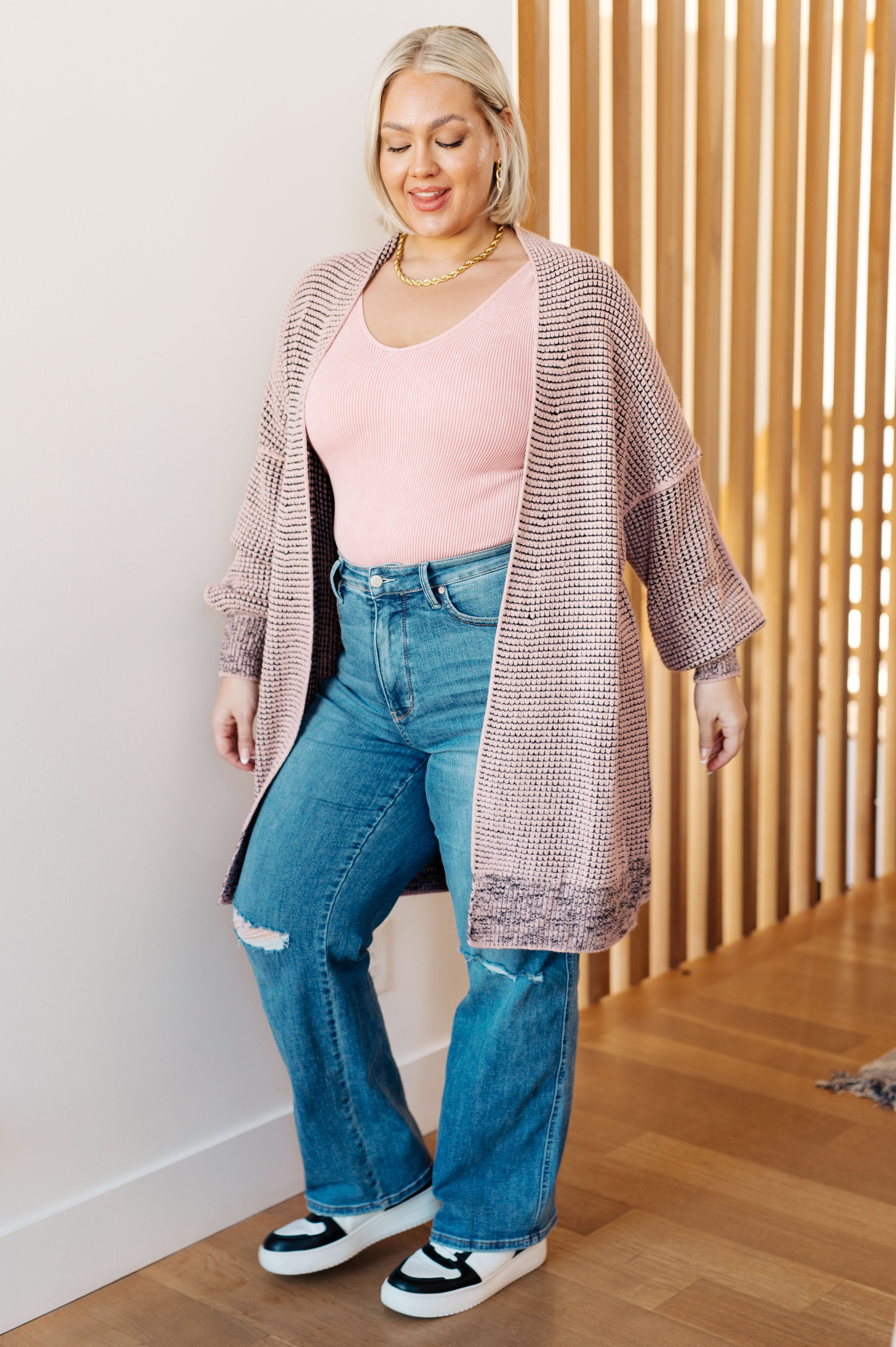 Curvy woman wearing mauve knit cardigan over pink tank top and distressed blue jeans standing indoors
