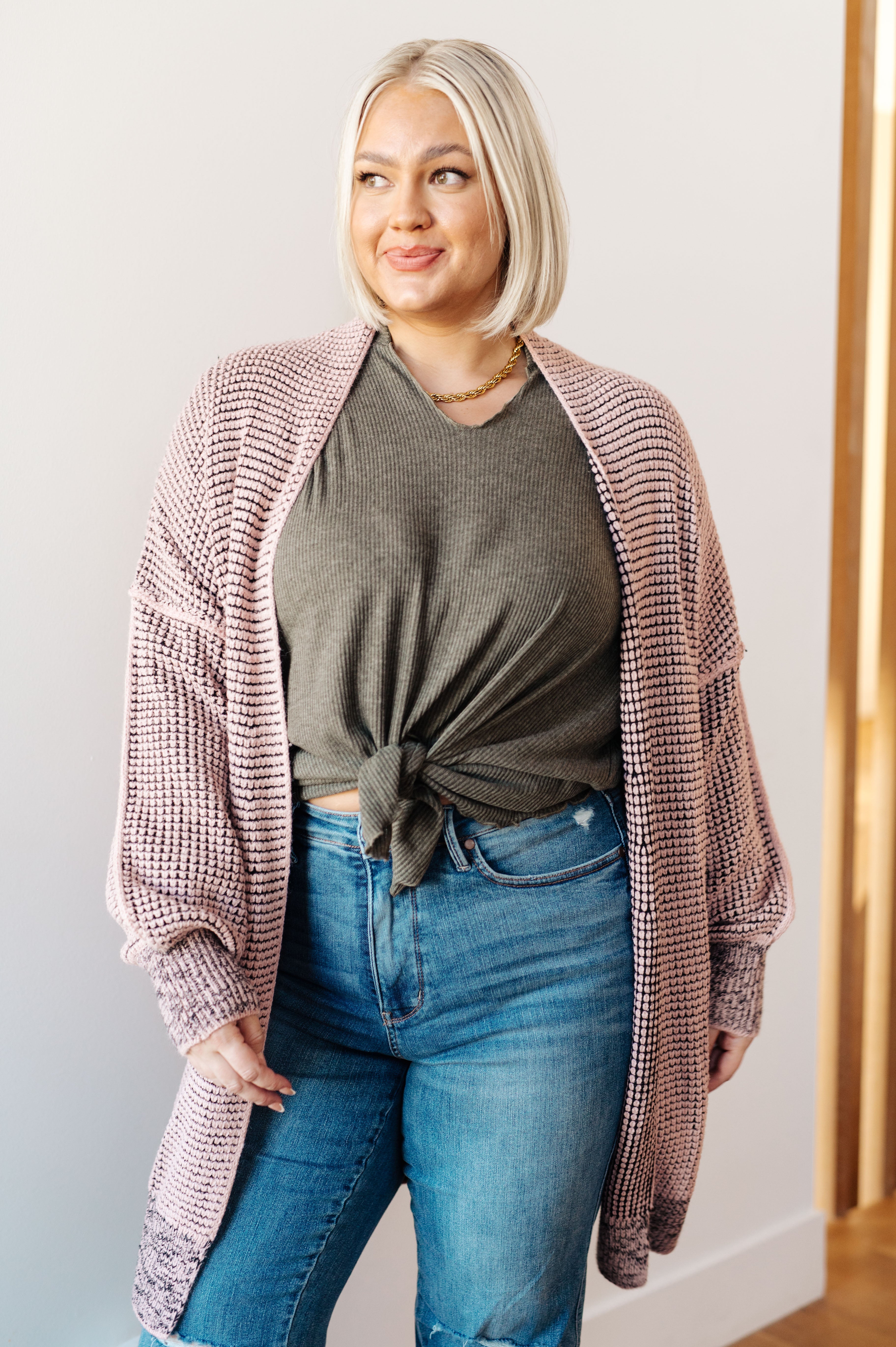 Woman wearing a mauve chunky knit cardigan over an olive green knotted top and blue distressed jeans indoors