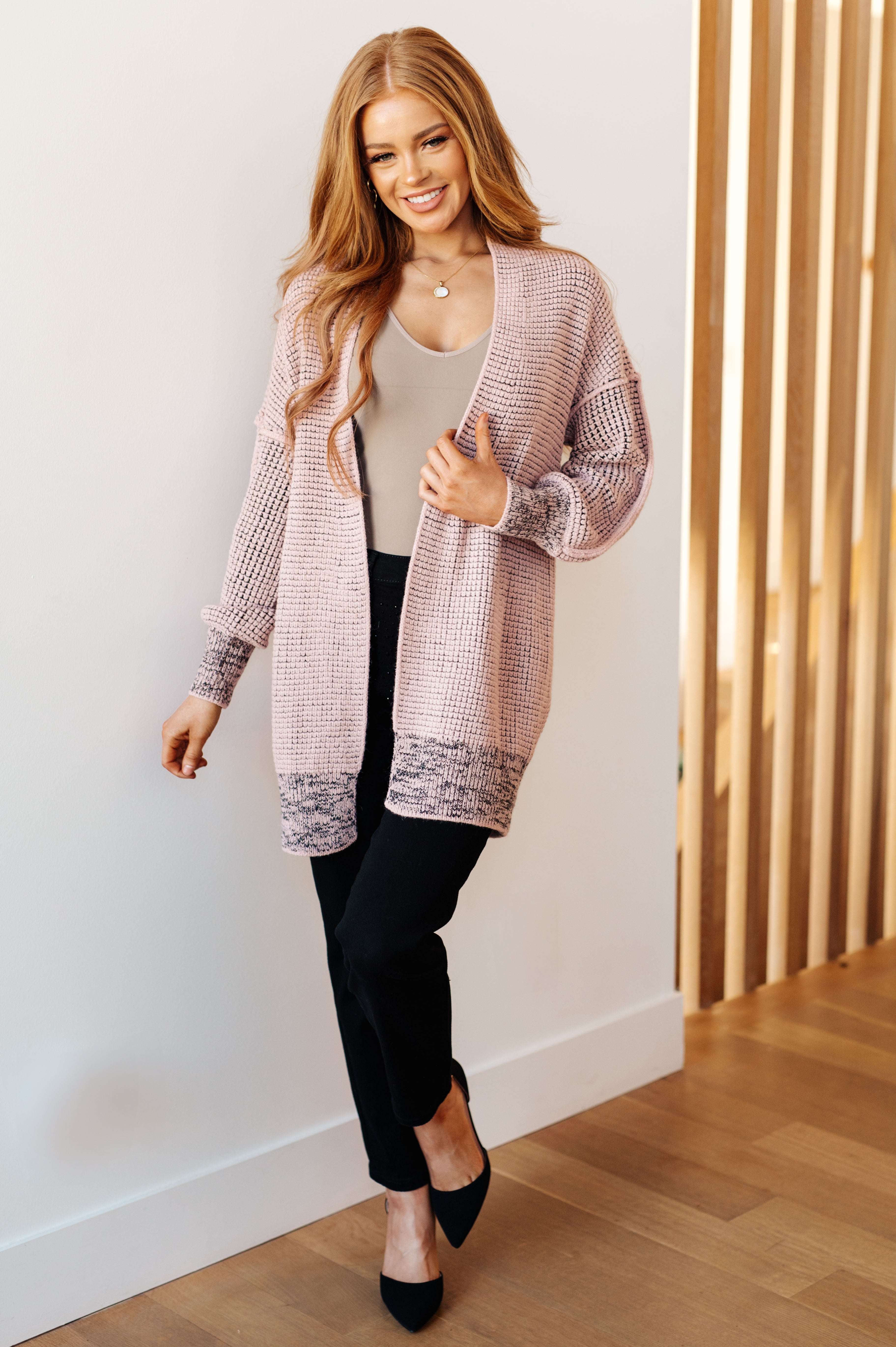 Woman wearing a mauve knit cardigan over a beige top and black pants, standing on wooden floor by slatted wall