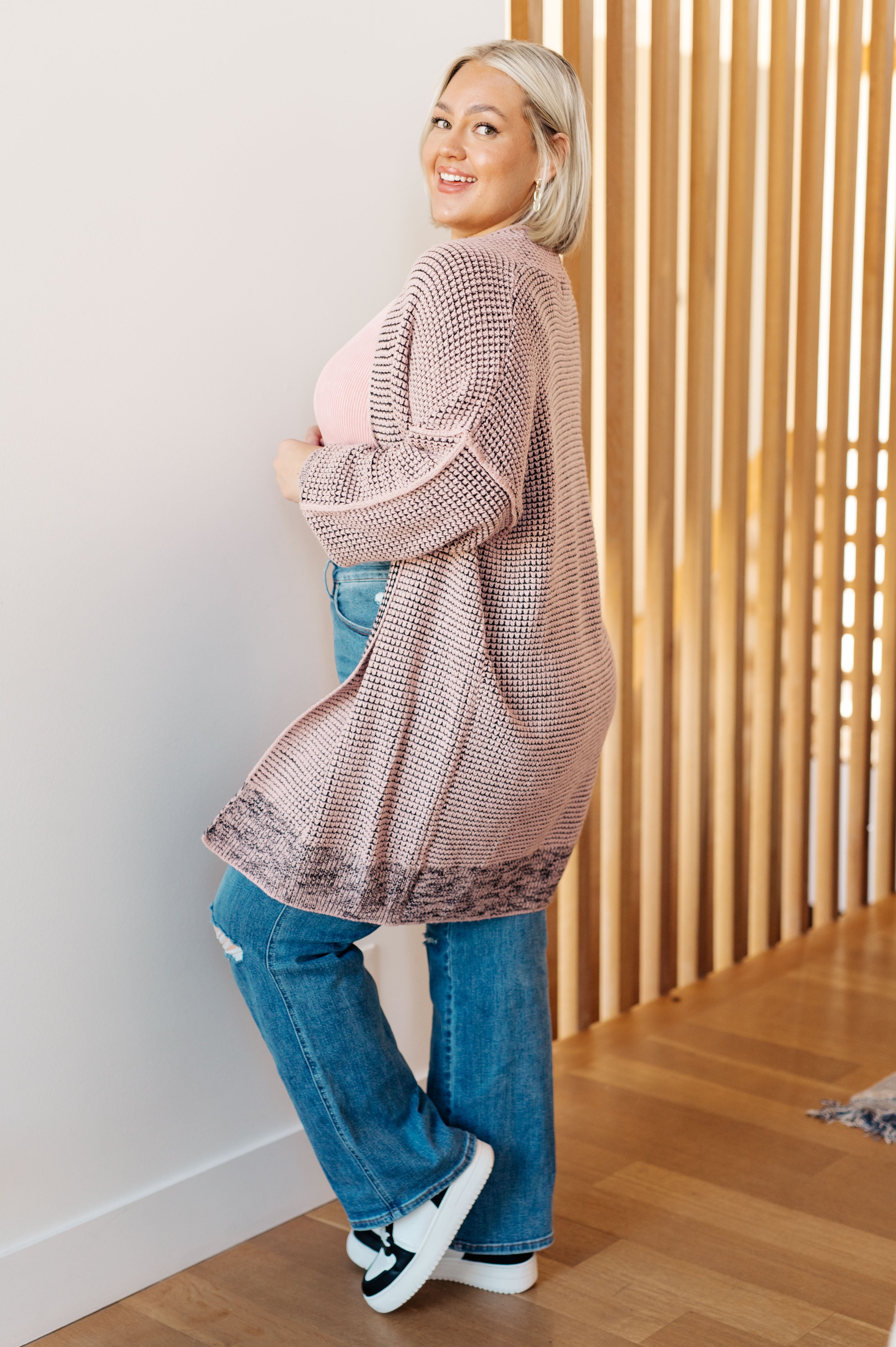 Smiling woman wearing a mauve knit cardigan over pink top and jeans, standing beside wooden slat wall indoors