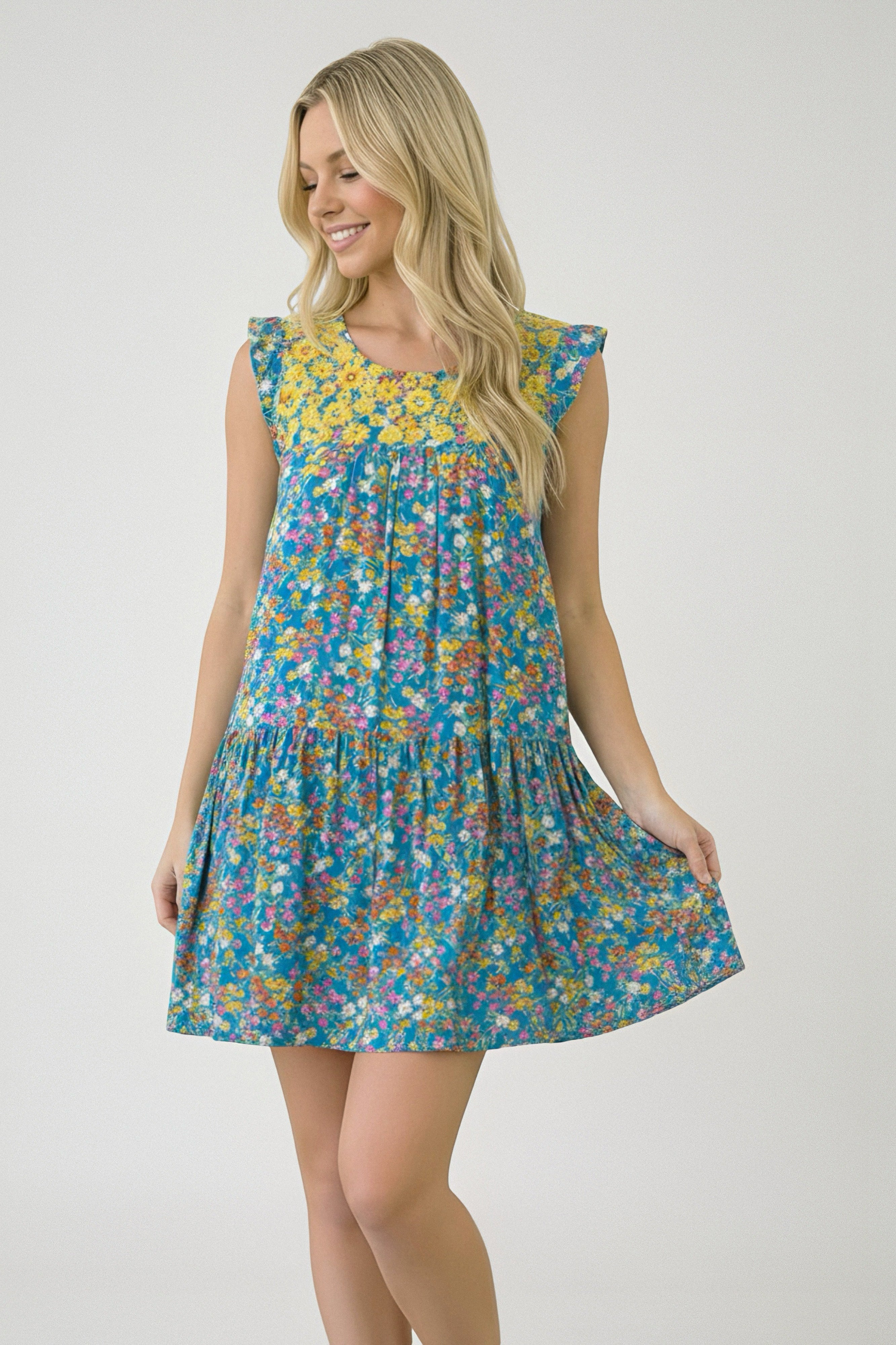 Smiling woman with long blonde hair wearing a blue floral dress with yellow yoke and flutter sleeves holding hem