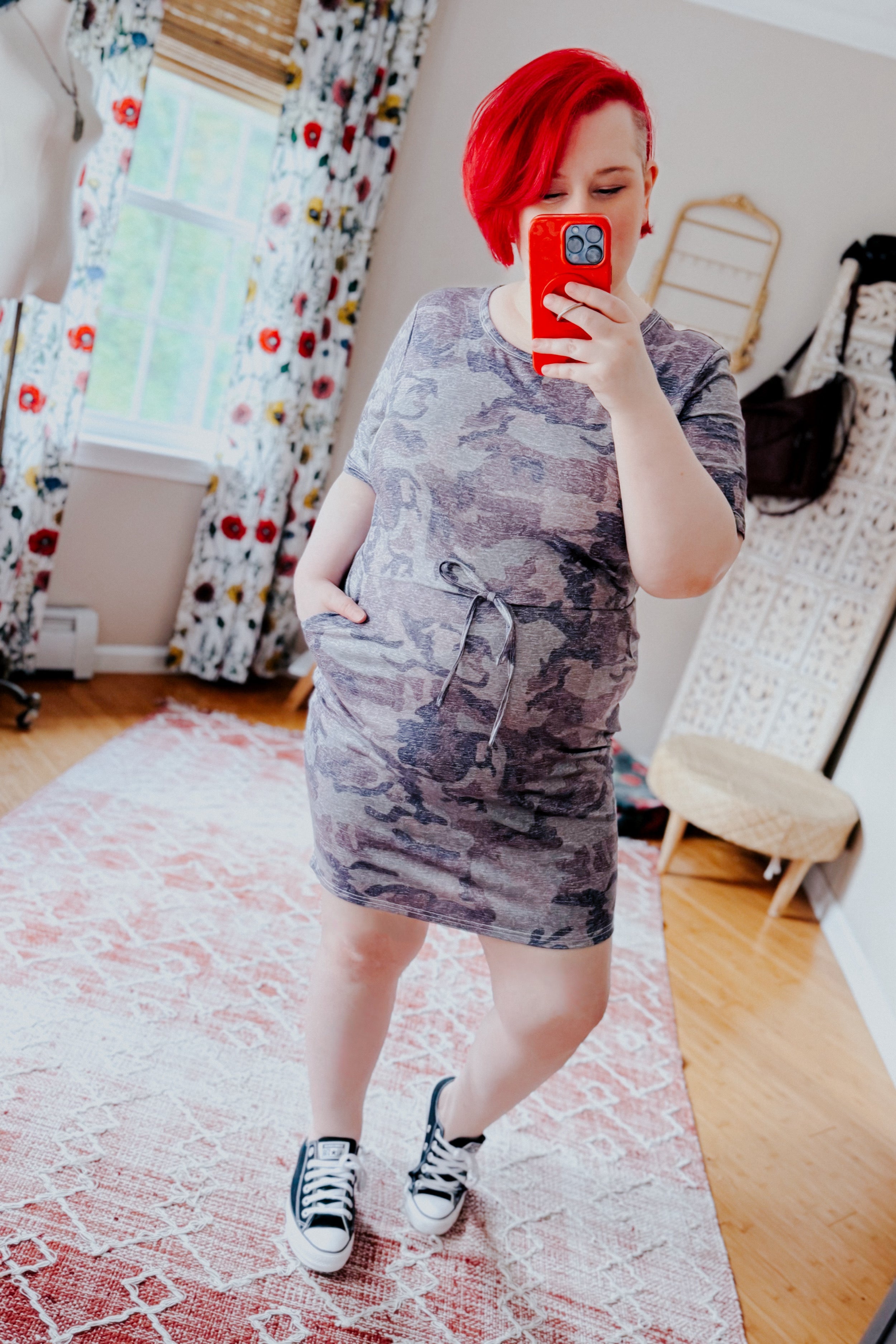 Woman with bright red hair wearing a blue camo print dress with drawstring waist and sneakers taking a mirror selfie indoors