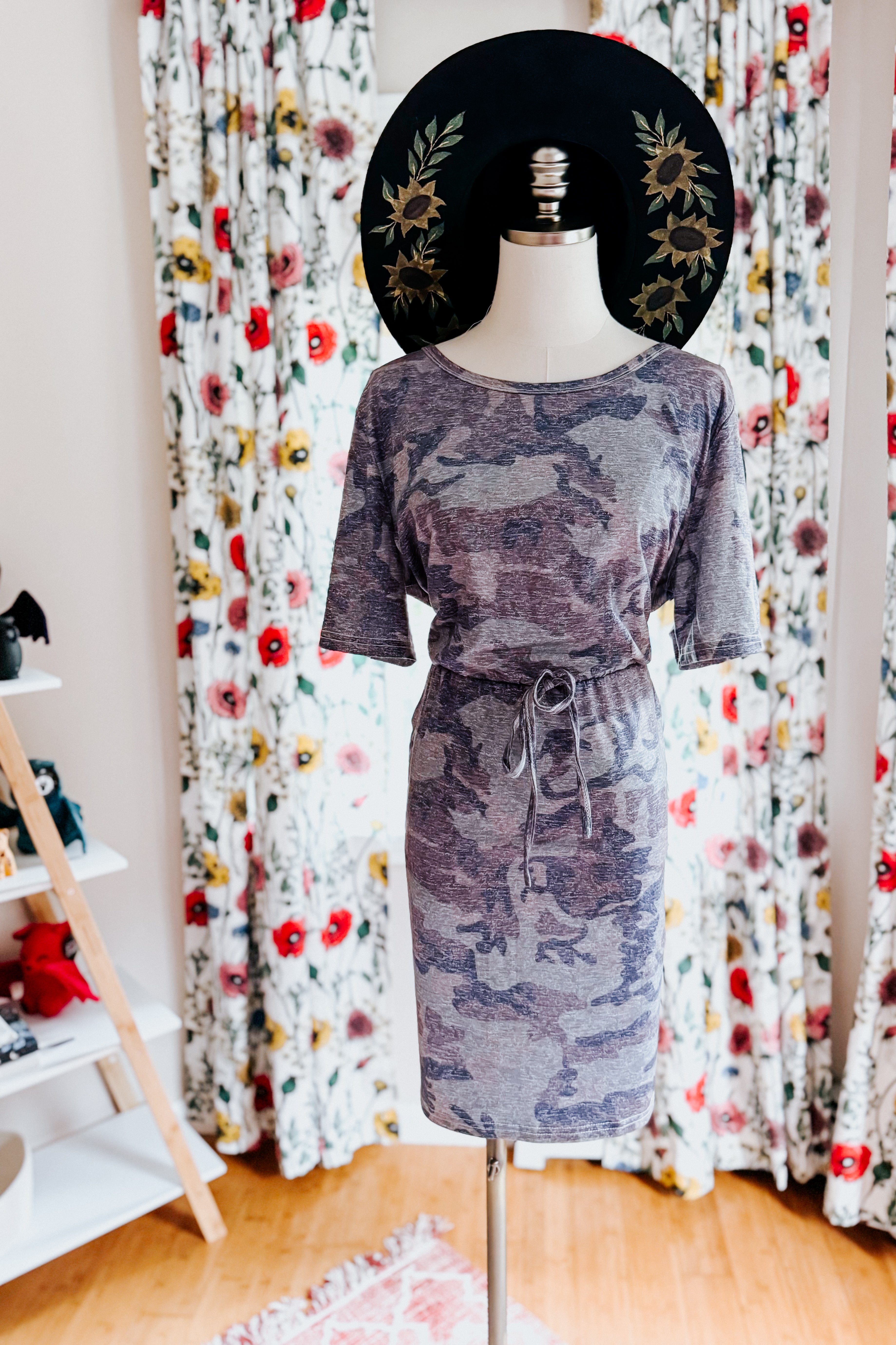 Mannequin dressed in a blue vintage camo print dress with a waist tie and a black hat featuring floral embroidery