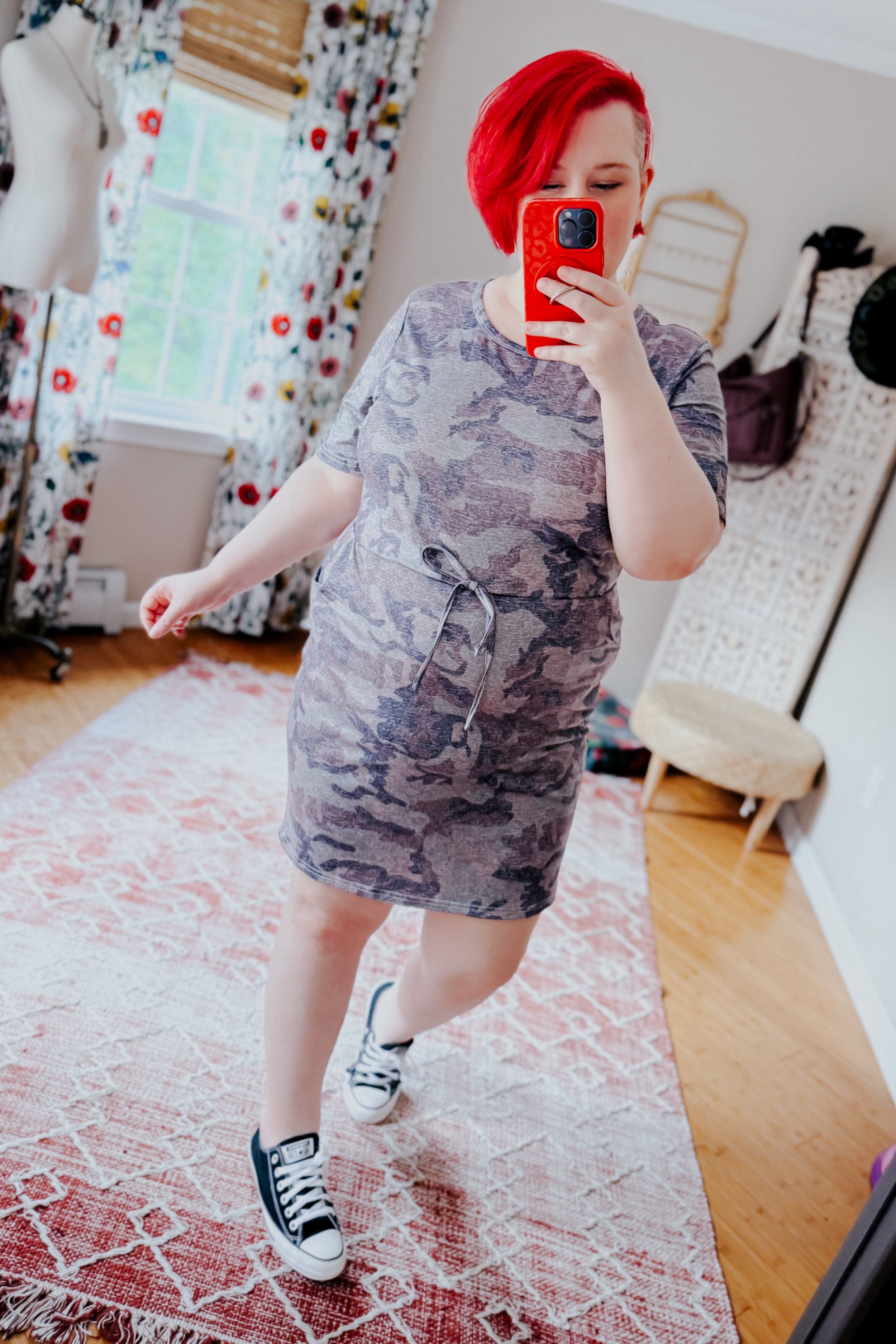 Woman wearing blue vintage camo print dress with tie waist, paired with black sneakers in bright indoor room