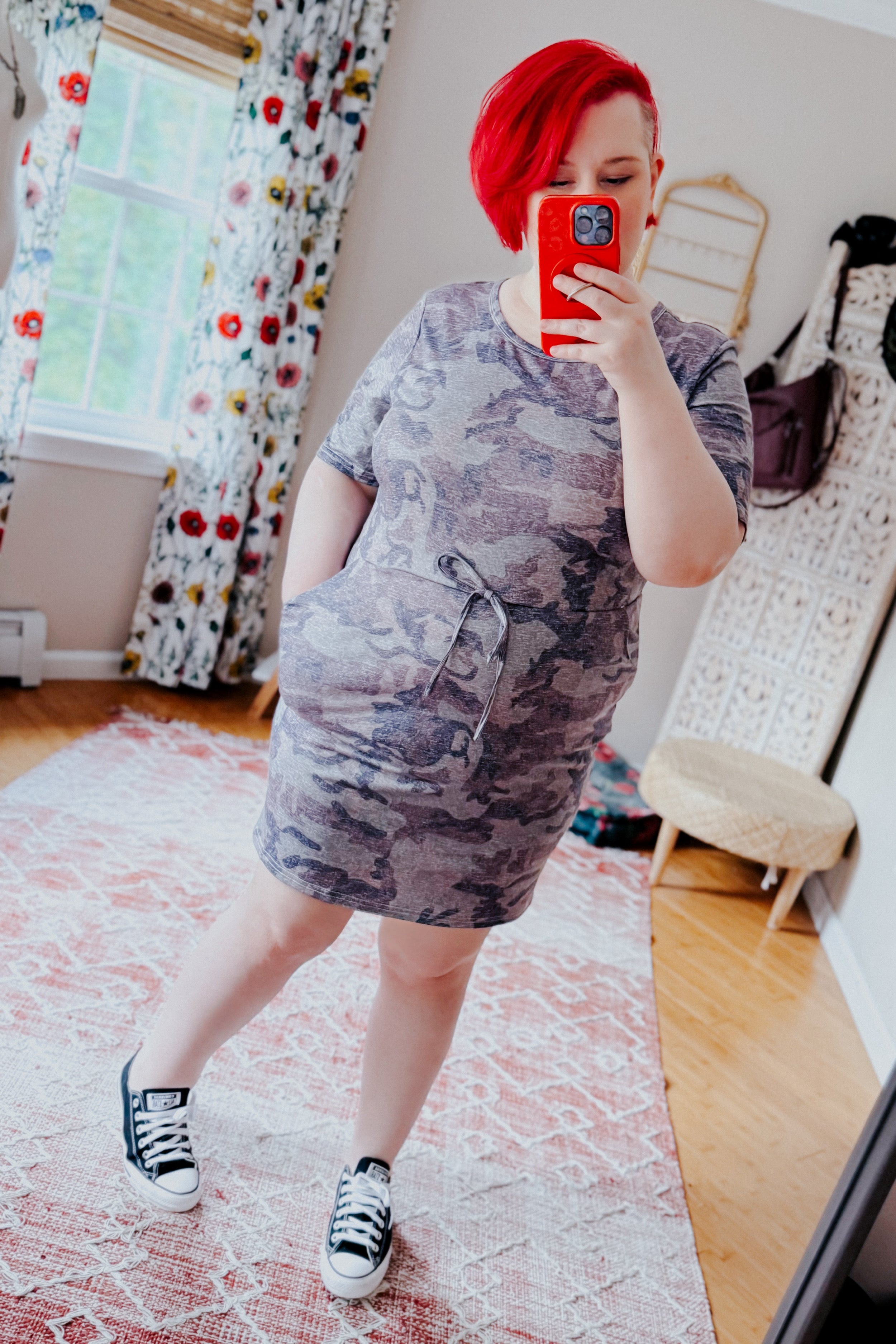 Woman with bright red hair wearing a blue vintage camo print dress with pockets and sneakers taking a mirror selfie indoors