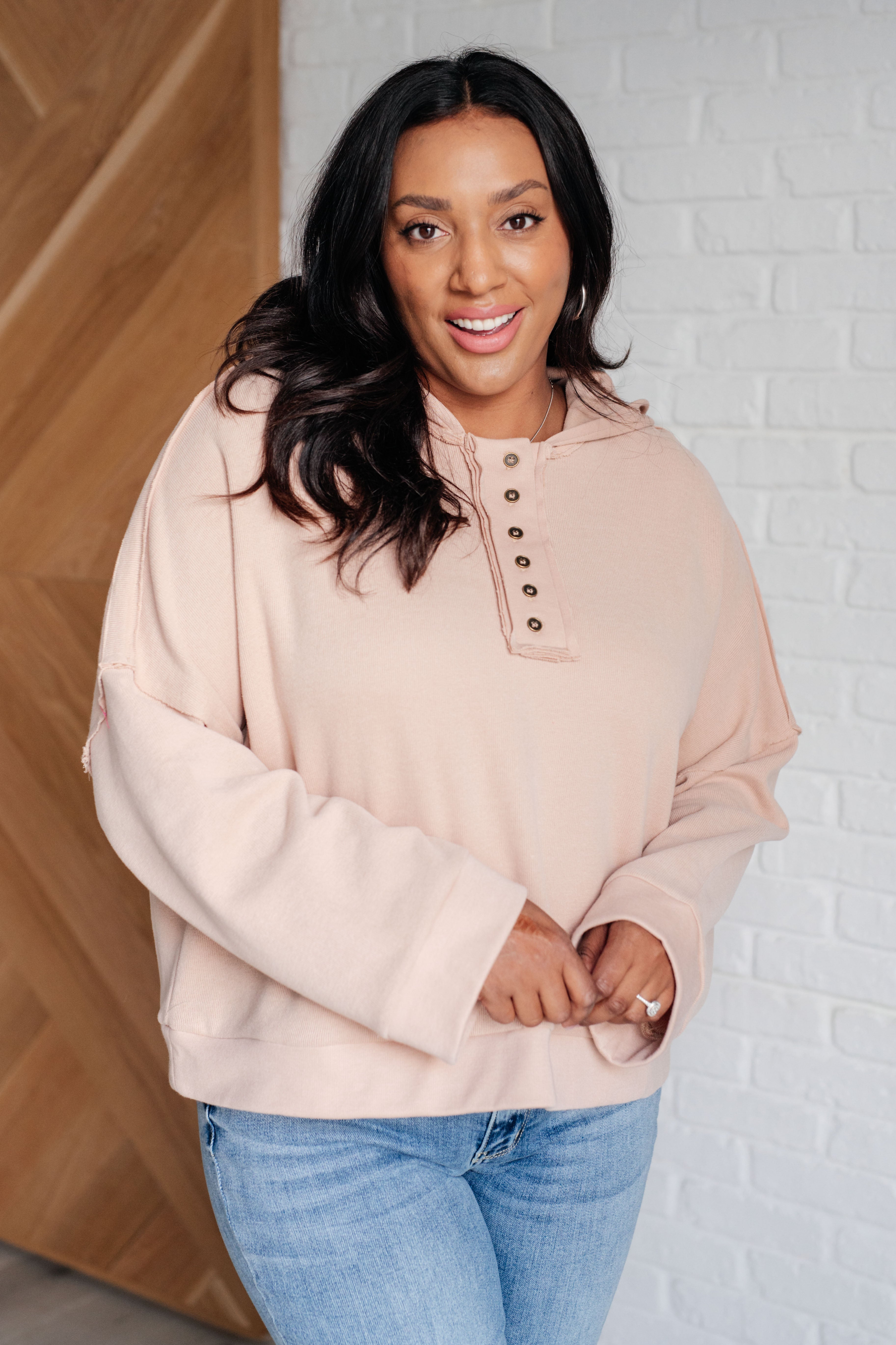 Plus-size model wearing a beige relaxed-fit hoodie with button details styled with light-wash jeans indoors