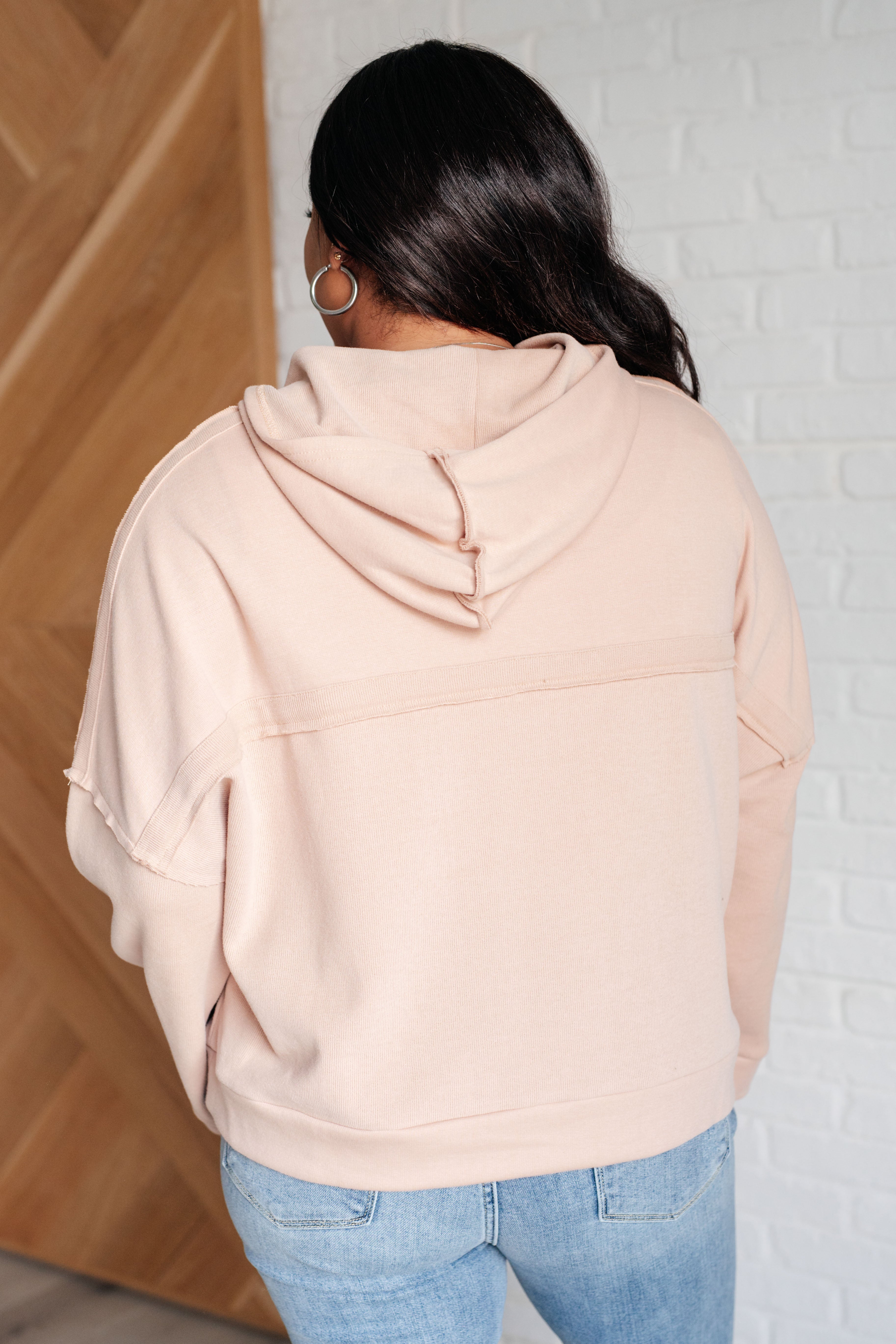 Woman wearing a beige oversized hoodie with hood up and hoop earrings, paired with light blue jeans indoors