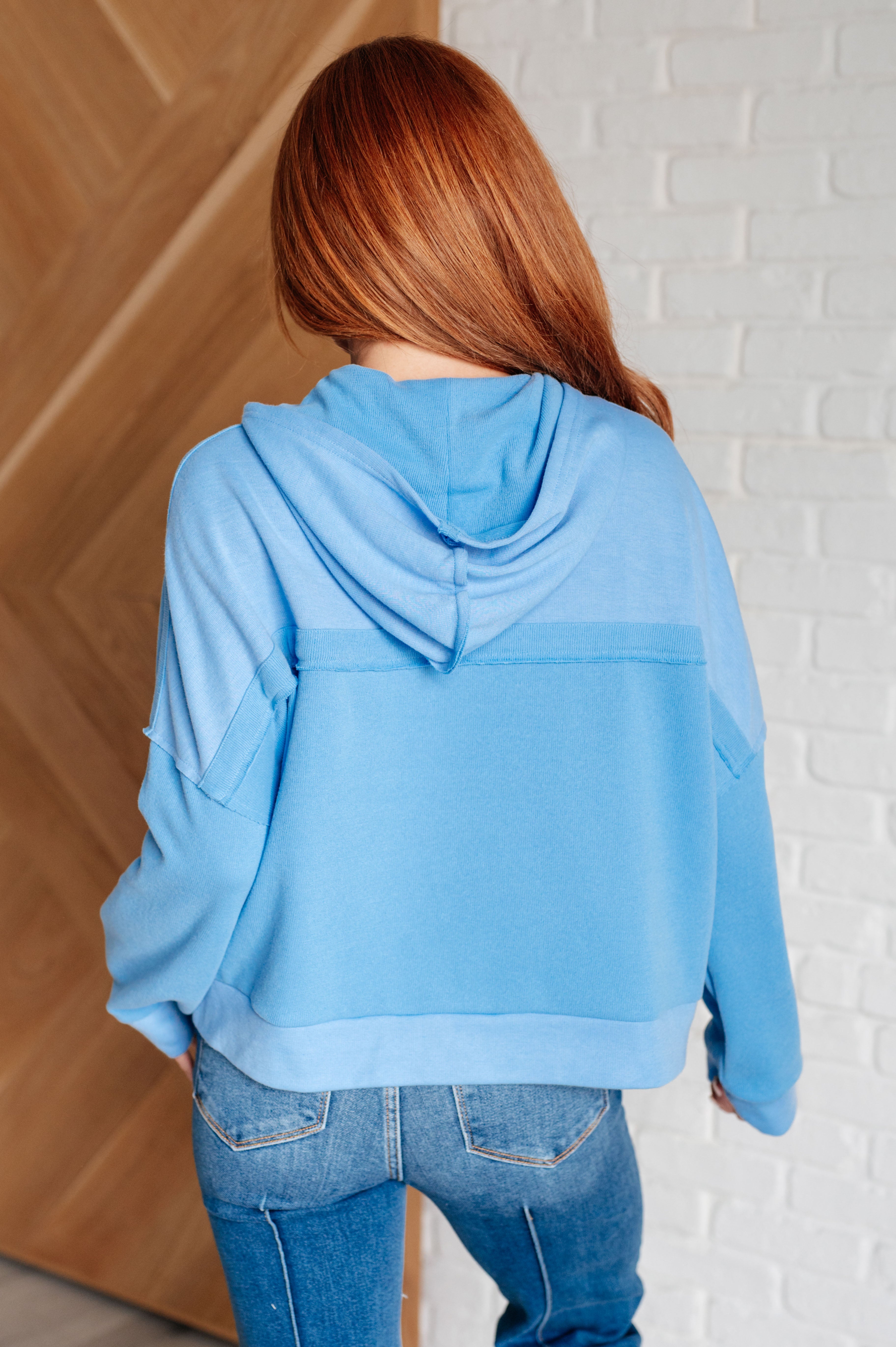 Woman with long red hair wearing a sky blue hooded sweatshirt paired with two-tone jeans indoors standing with back facing camera